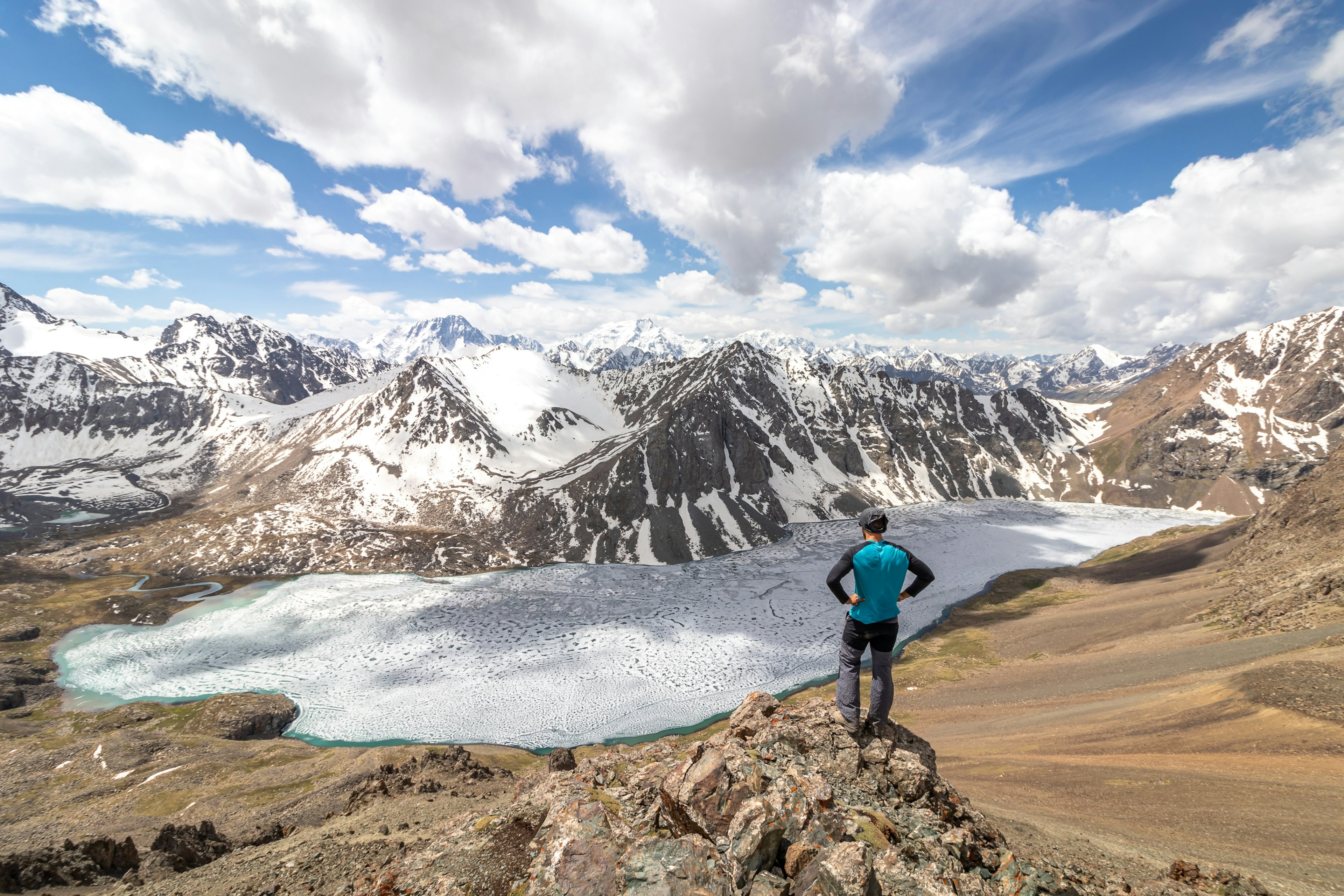 A man stands on rocks and looks down at a frozen mountain lake. The ground is brown and dry, and snow-covered mountains rise from the other side of the lake.