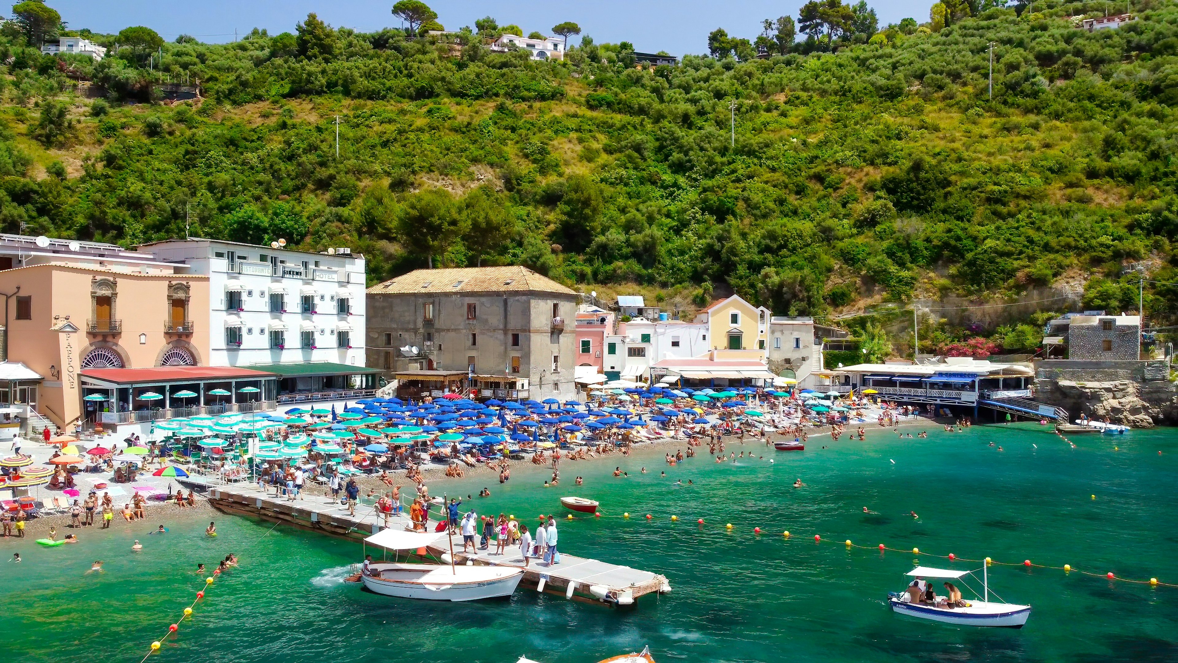 Swimmers, day trippers, and sunbathers fill the sea, harbor and beach in a small town on a sunny day