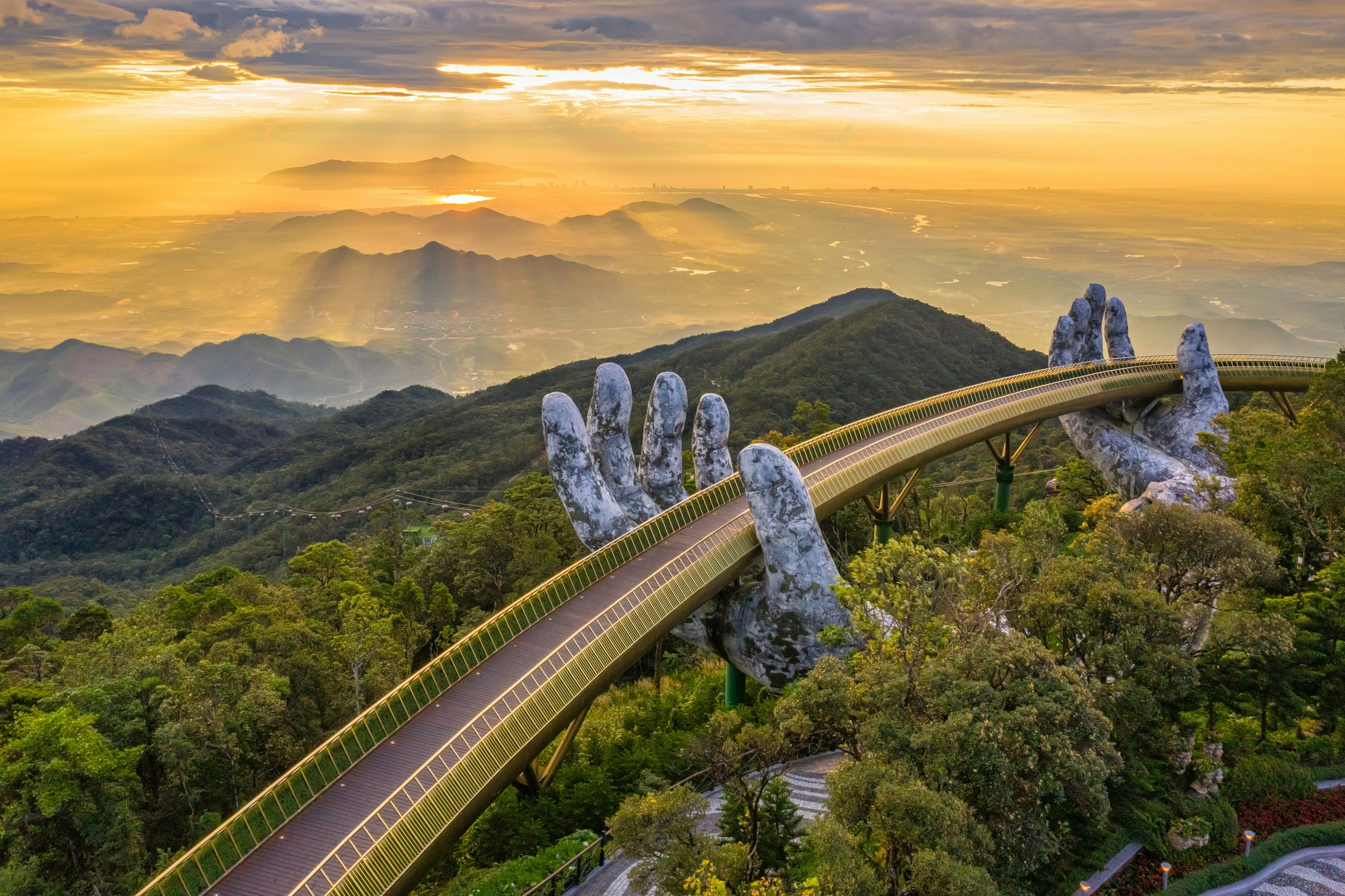 A pedestrian bridge in a mountainous area at sunset that appears to be supported by two concrete hands