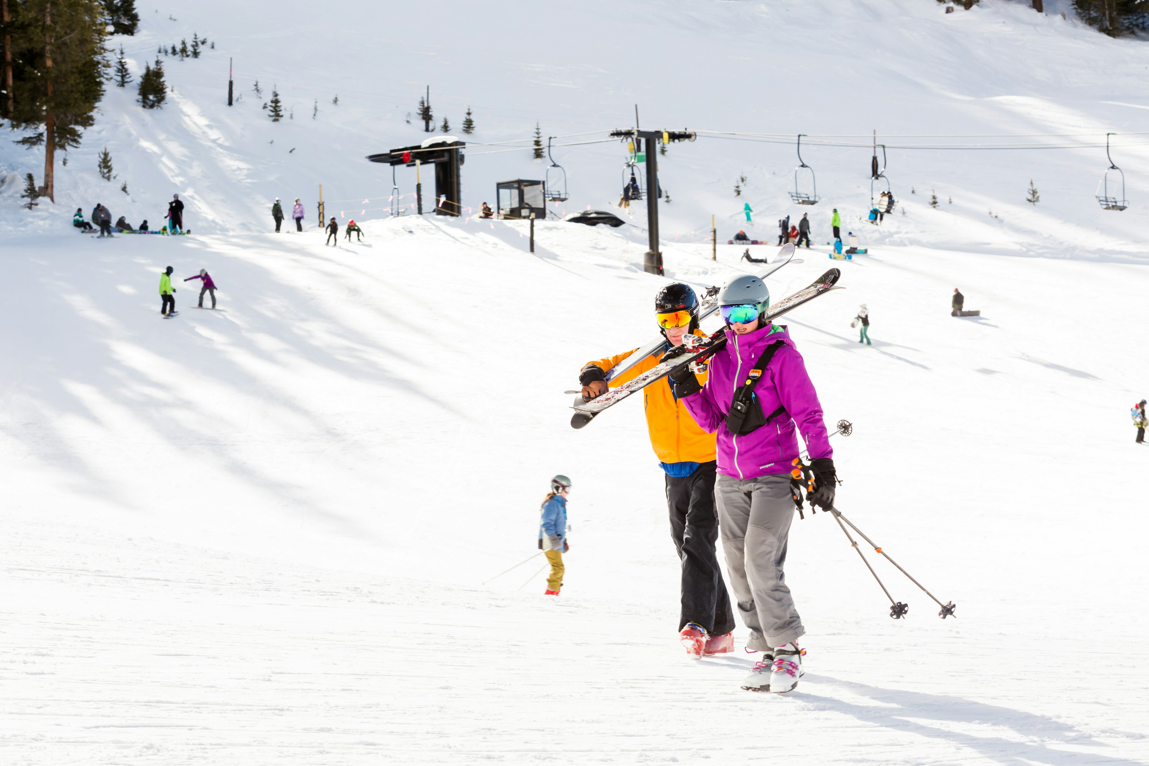 Two skiers wearing bright colors carry their skis at the bottom of a slope, with a ski lift in the background.