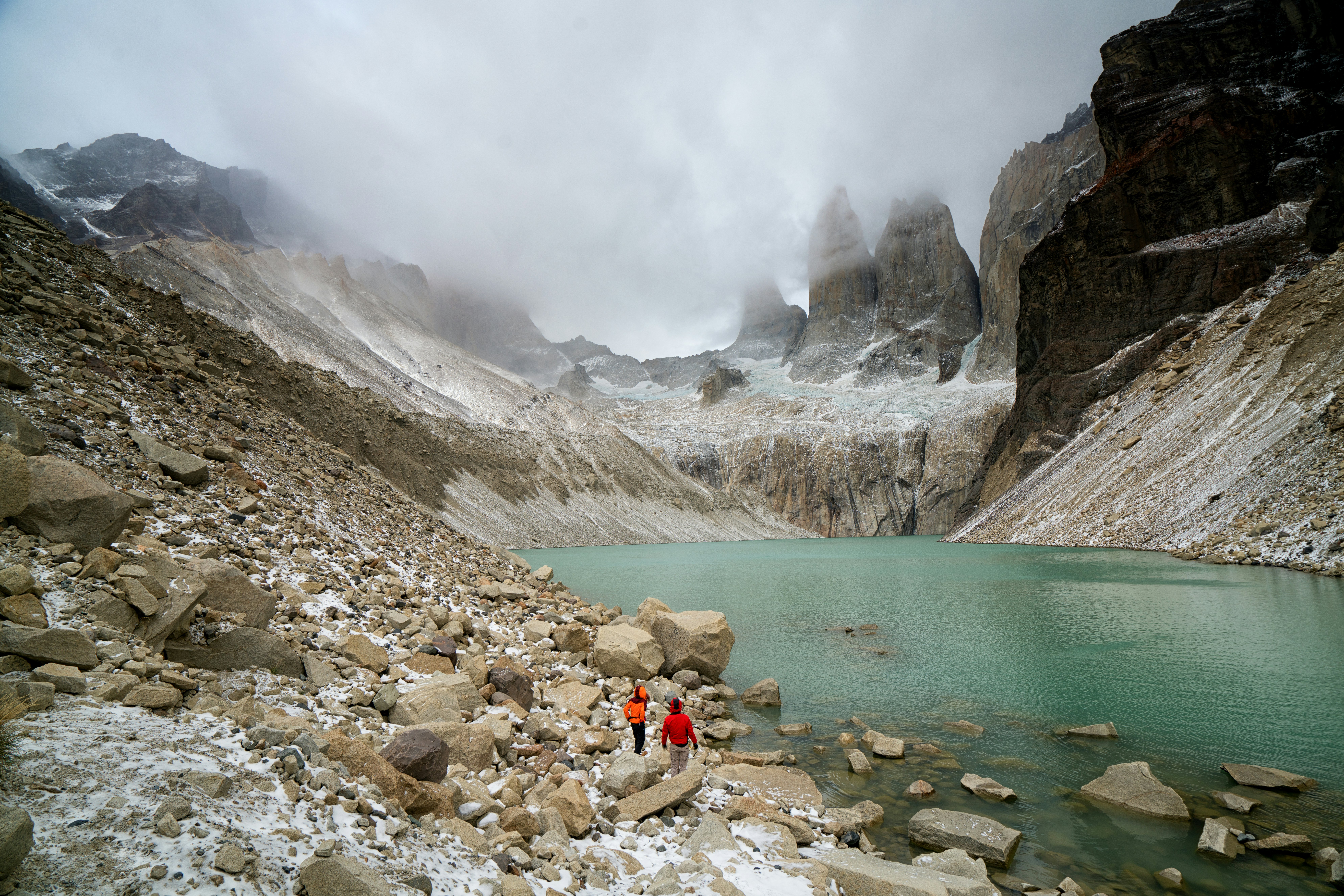 Two people in red jackets walk on a rocky hillside toward a clear blue lake surrounded by mountains; a low cloud hangs in the valley at the far shore.