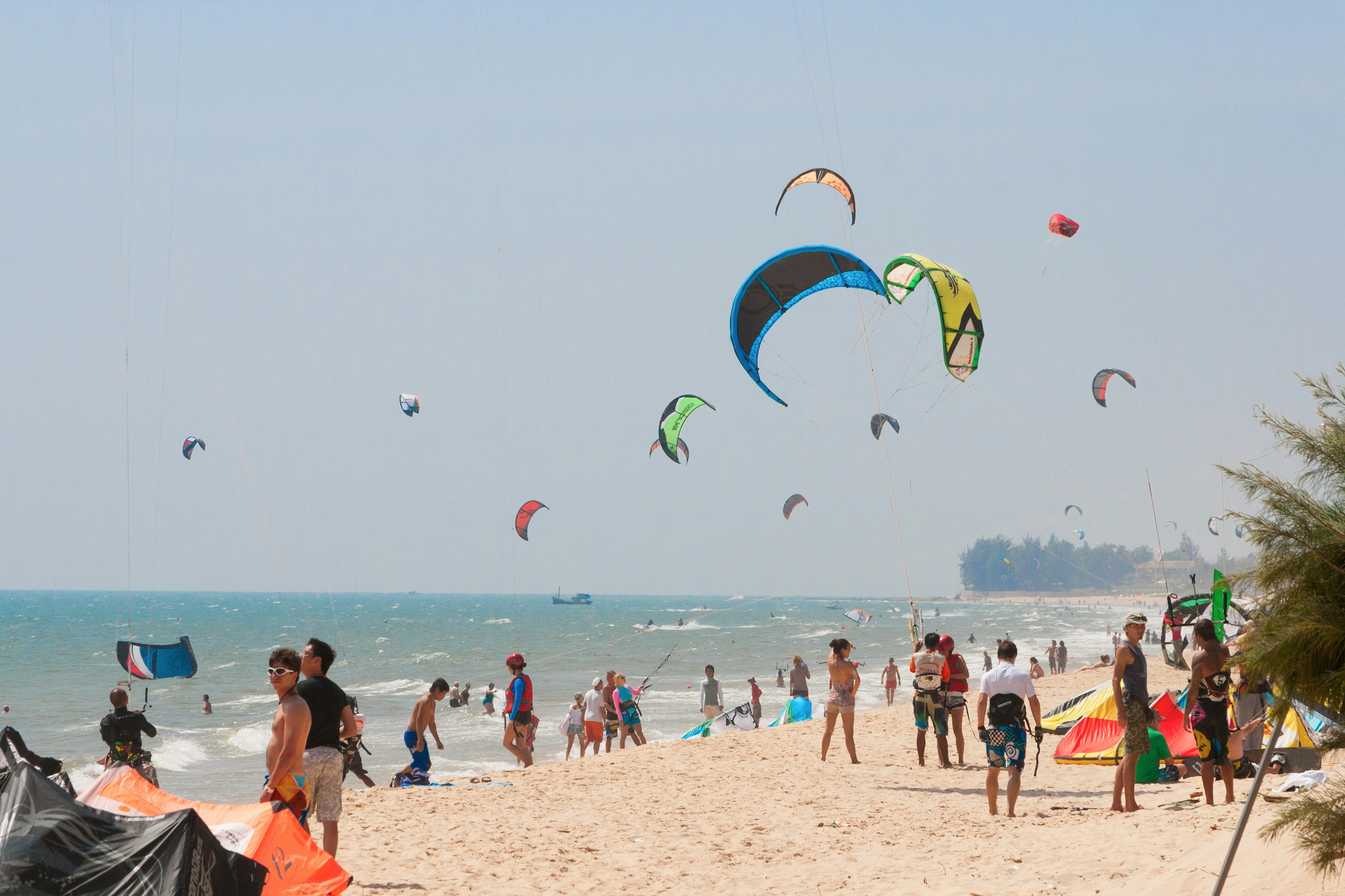 Mui Ne beach in Vietnam. The beach is a strip of sand with sunbathers and people walking, while the sea is filled with kite surfers, whose colourful kites are visible in the sky above.