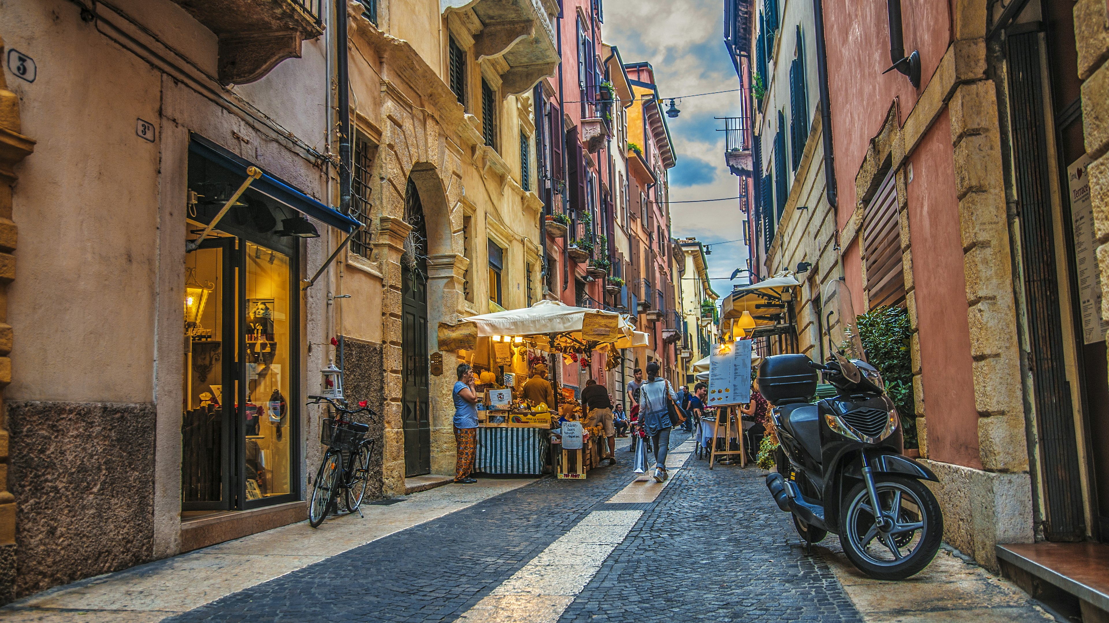 A narrow street with shops, a restaurant terrace and a fresh food stall