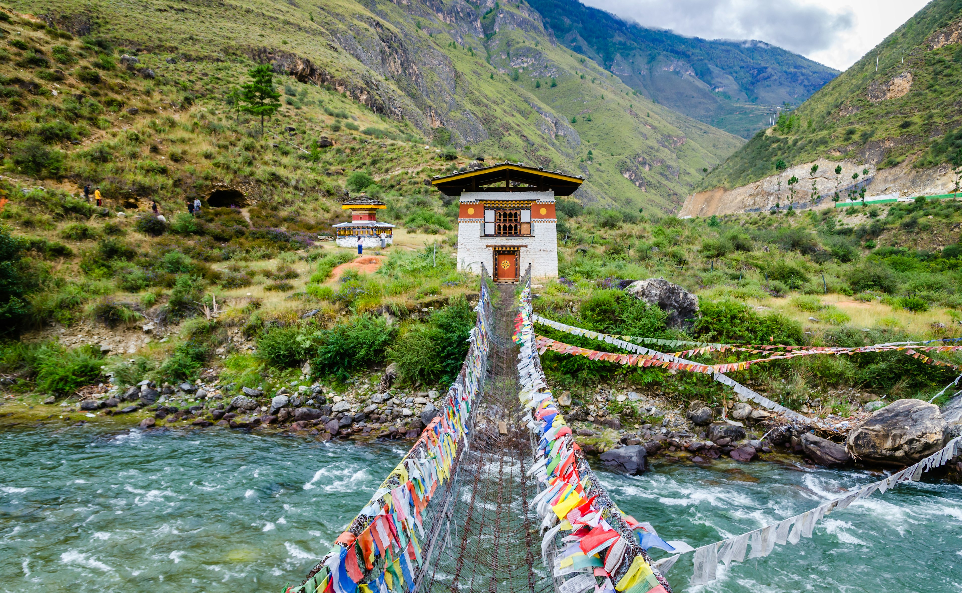 A hanging chain bridge lined with colorful flags crosses a fast-flowing river in a mountainous region