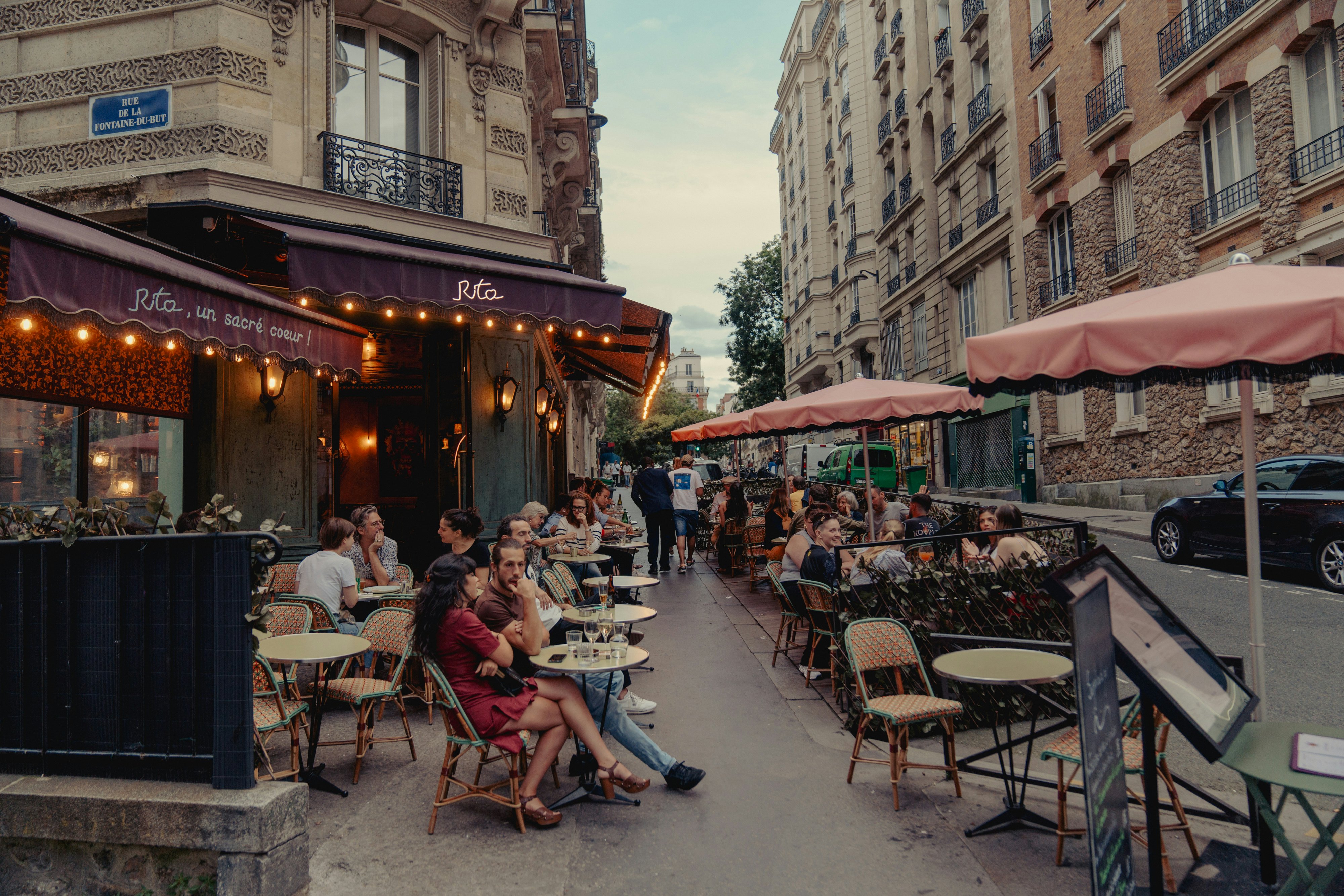 People sit at outdoor tables under a red awning with lights on a street in the 18th arrondissement of Paris, France