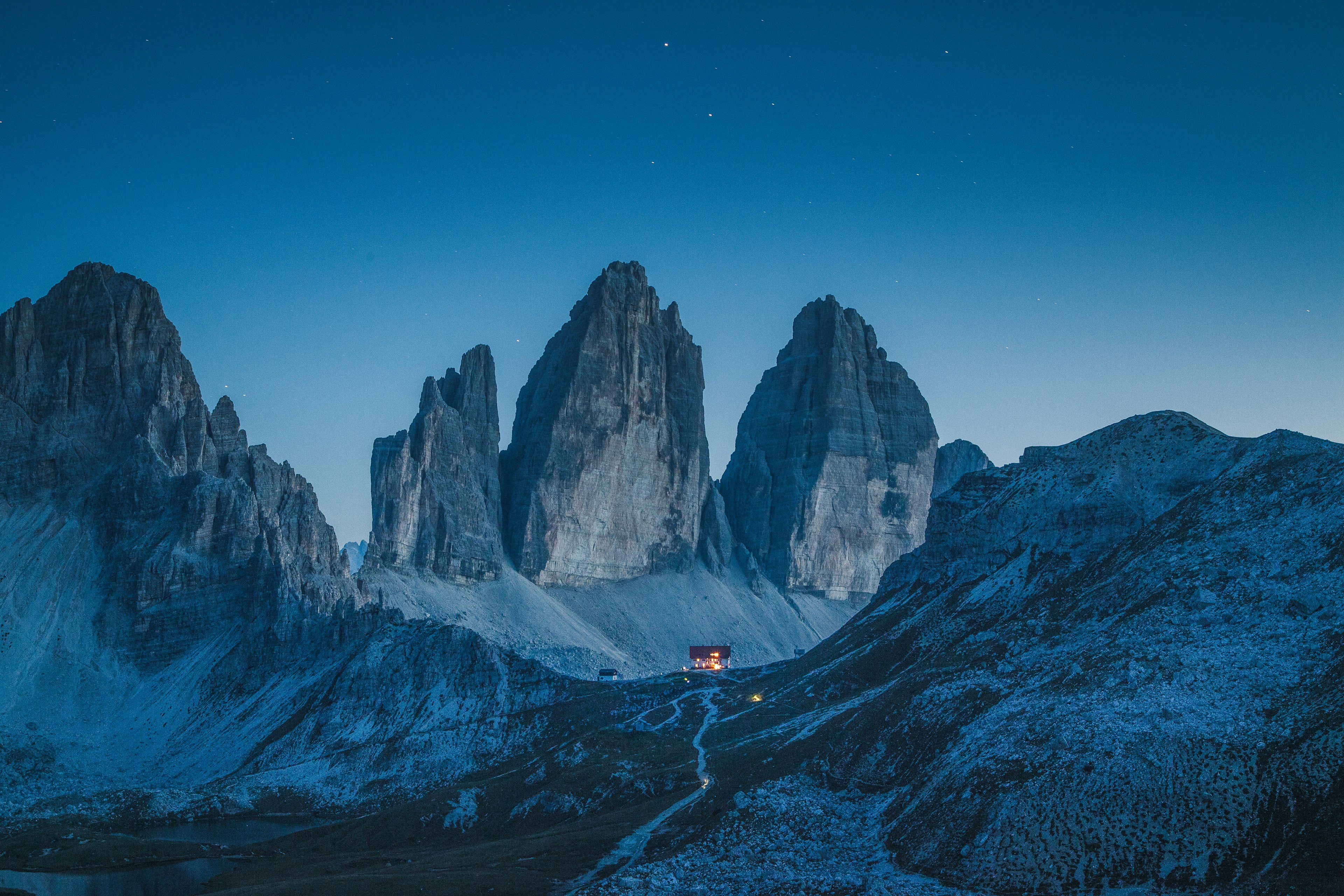 A wide shot of a hut at night, illuminated from within, surrounded by dramatic, snow-covered limestone mountain peaks