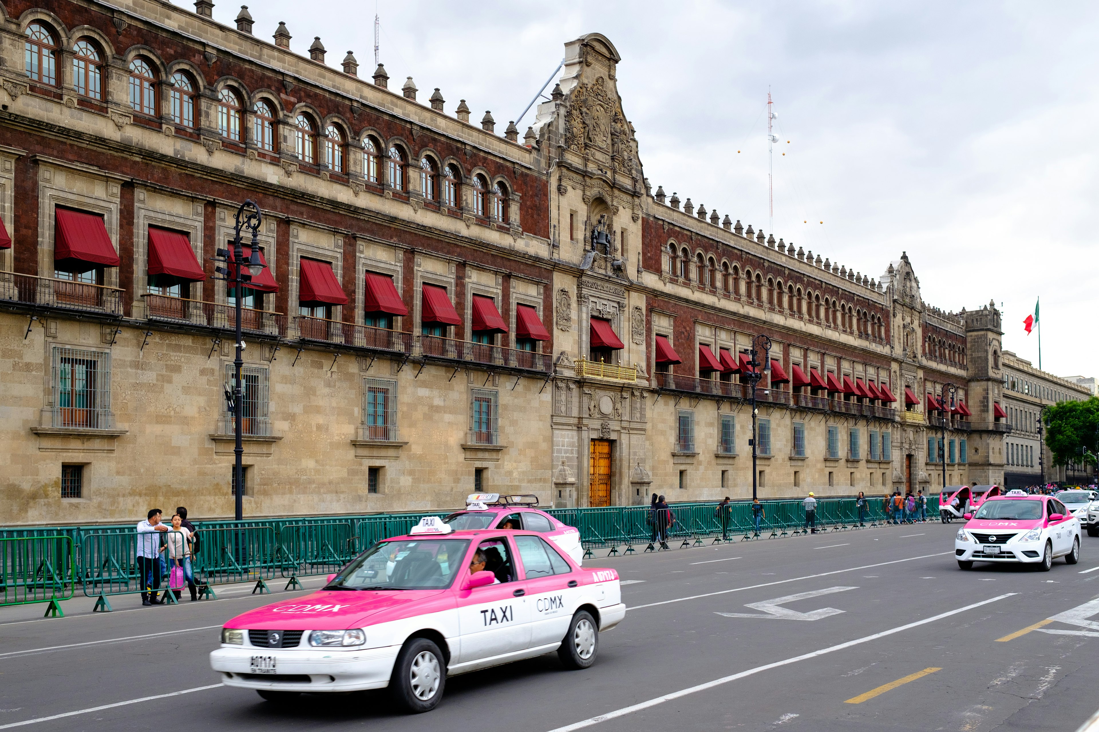 Pink-and-white taxis drive down a street next to an imposing building with red awnings and crenellations