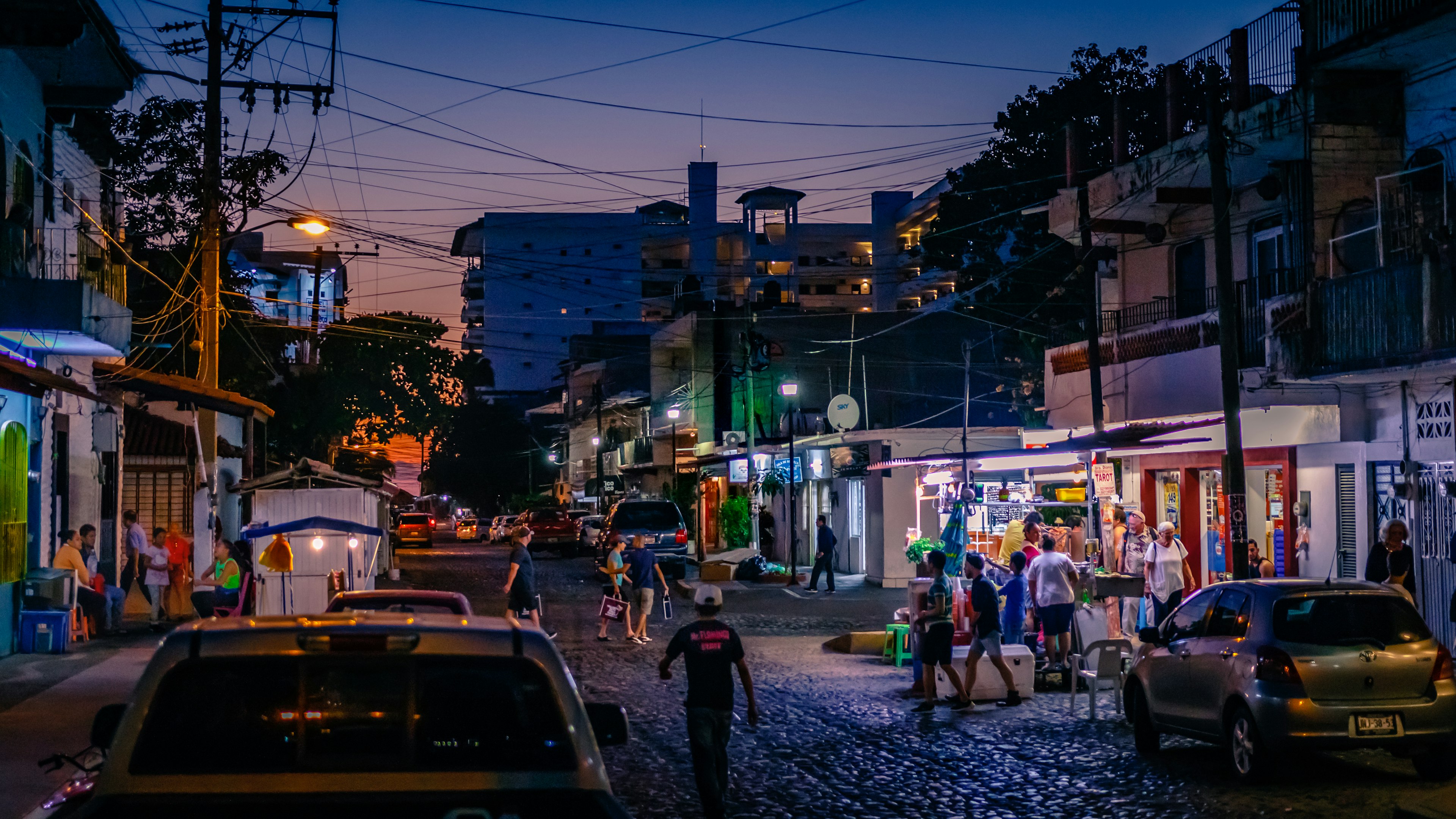 People walk at sunset on a cobblestone street in a city, with the lights of restaurants and businesses illuminating the scene