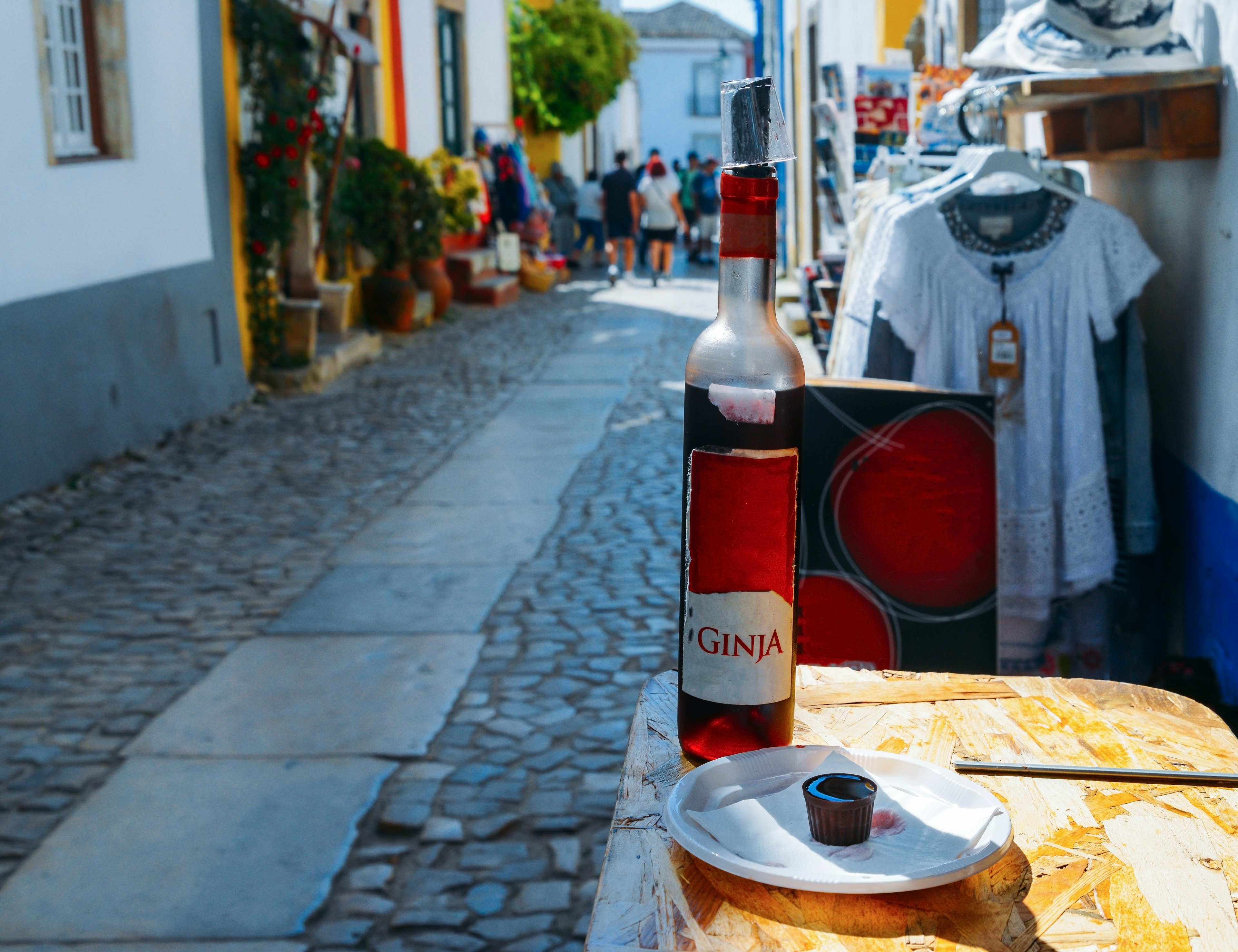 A cobbled street in a medieval town lined with small shops. A bottle of cherry liqueur called "ginja" stands on display next to a cup made of chocolate