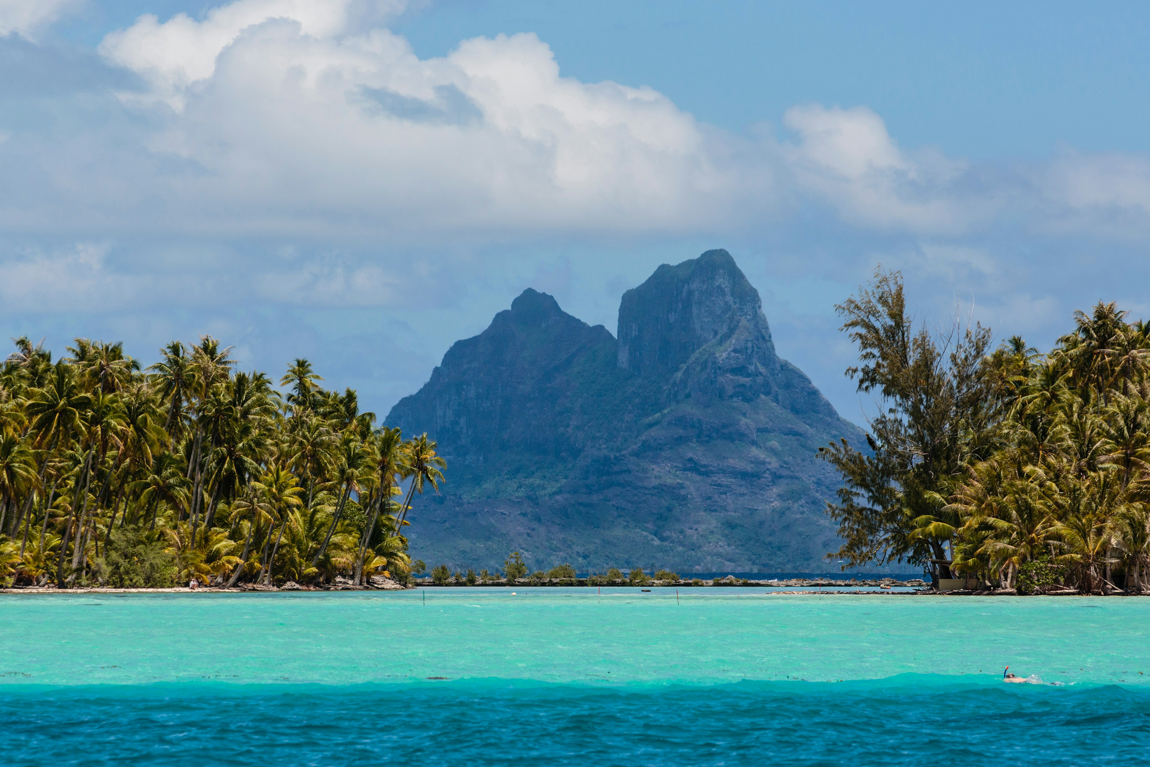 A double-peaked mountain is visible in the distance behind bright blue waters fringed with dense palms