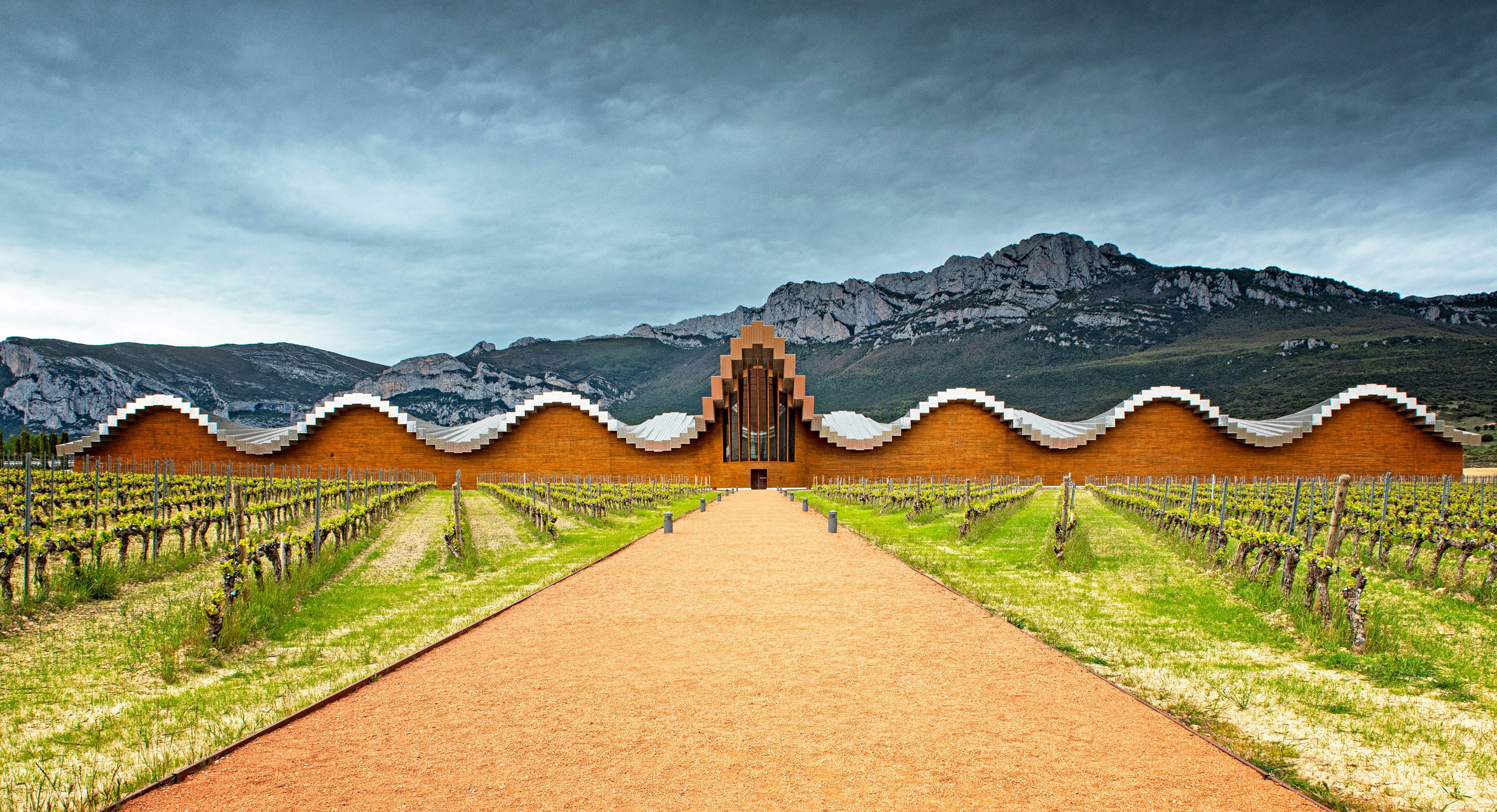 A dirt path between rows of low green shrubbery leads to a structure with a metallic sloping roof and a wooden entrance. A large mountain can be seen in the distance