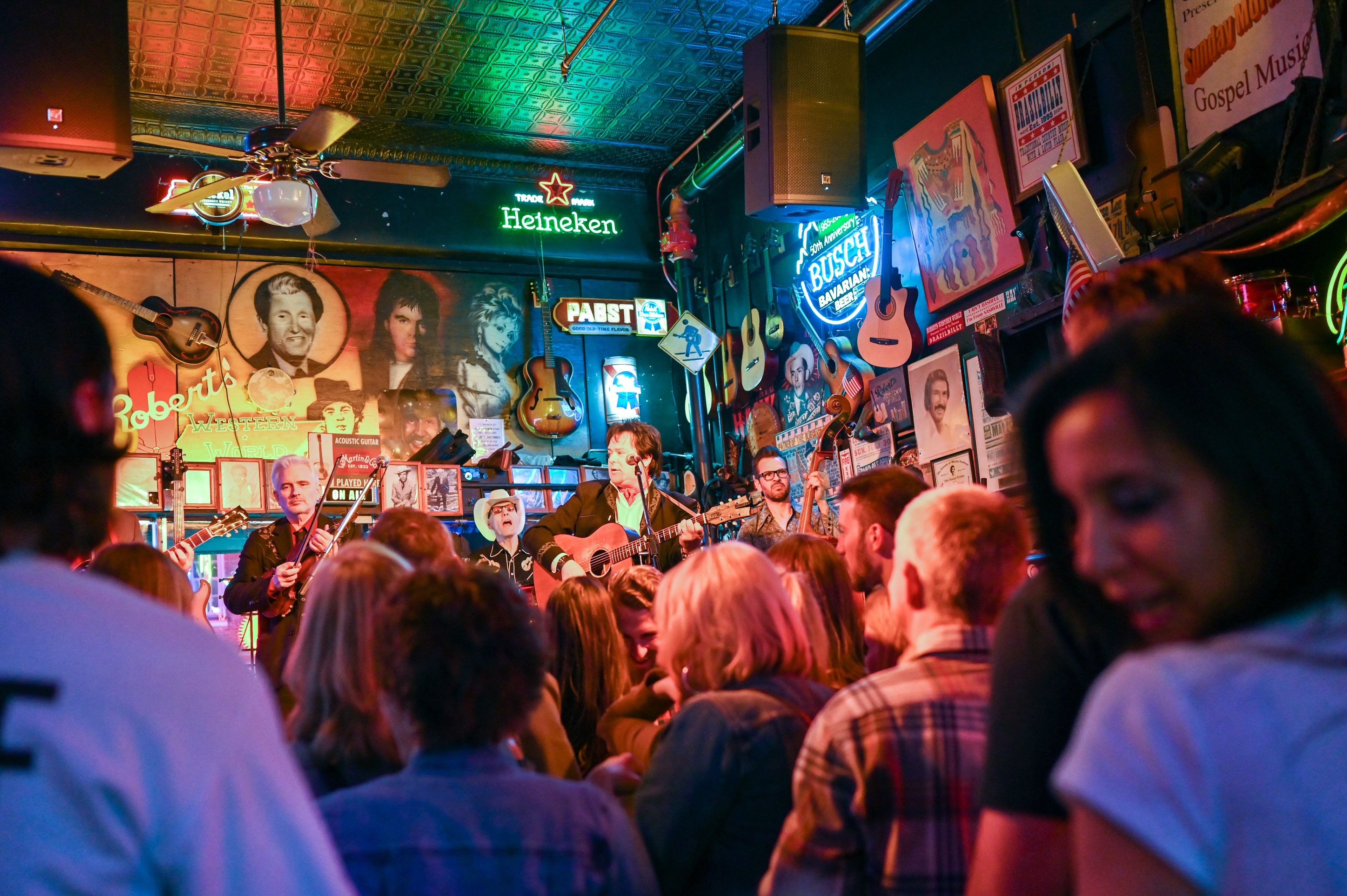 A crowd listens to a live band perform in a crowded venue with neon signs and vintage posters on the walls