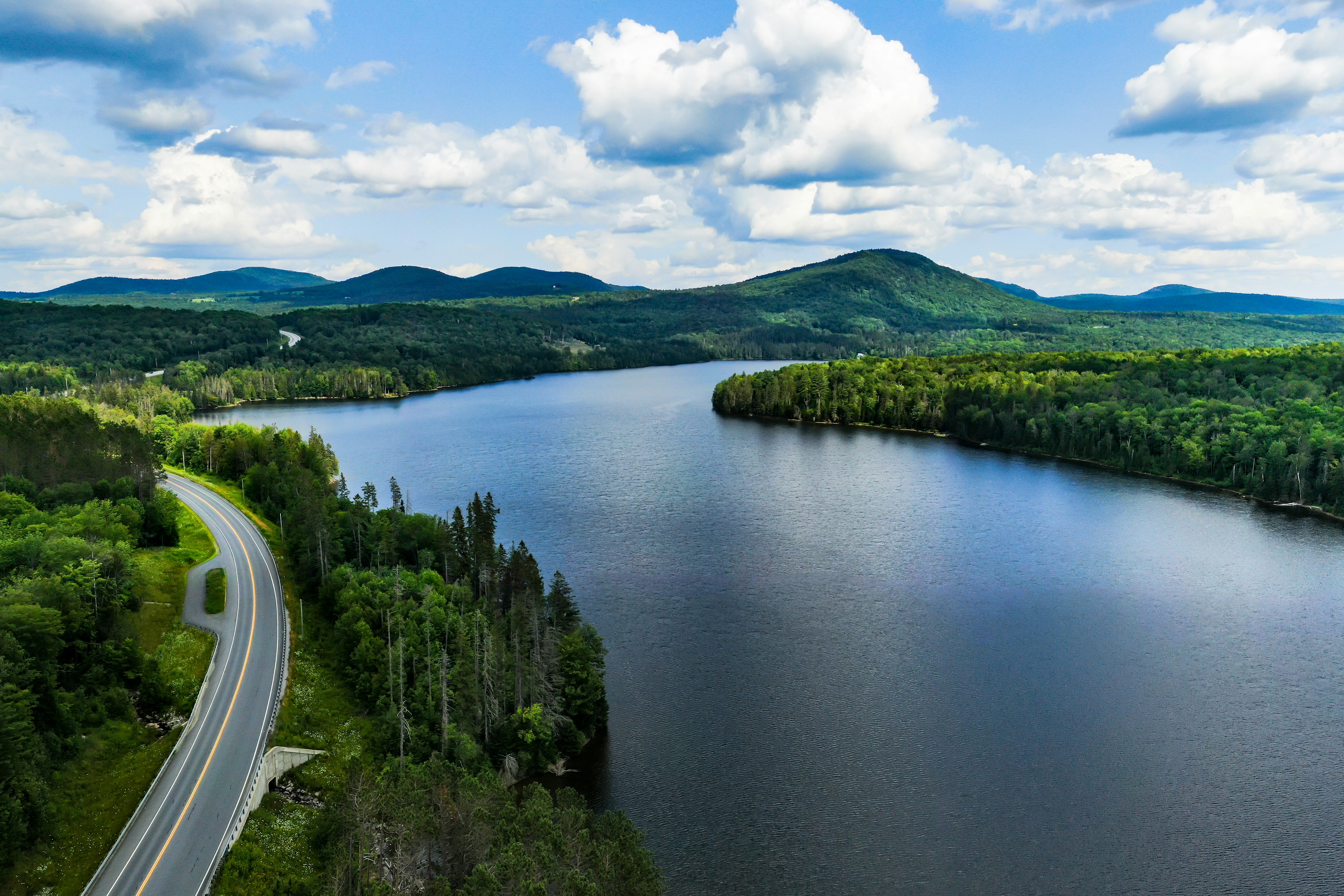 An aerial shot of a lake with a road along its shore, under puffy clouds in the sky. Green trees and hills are visible in the distance