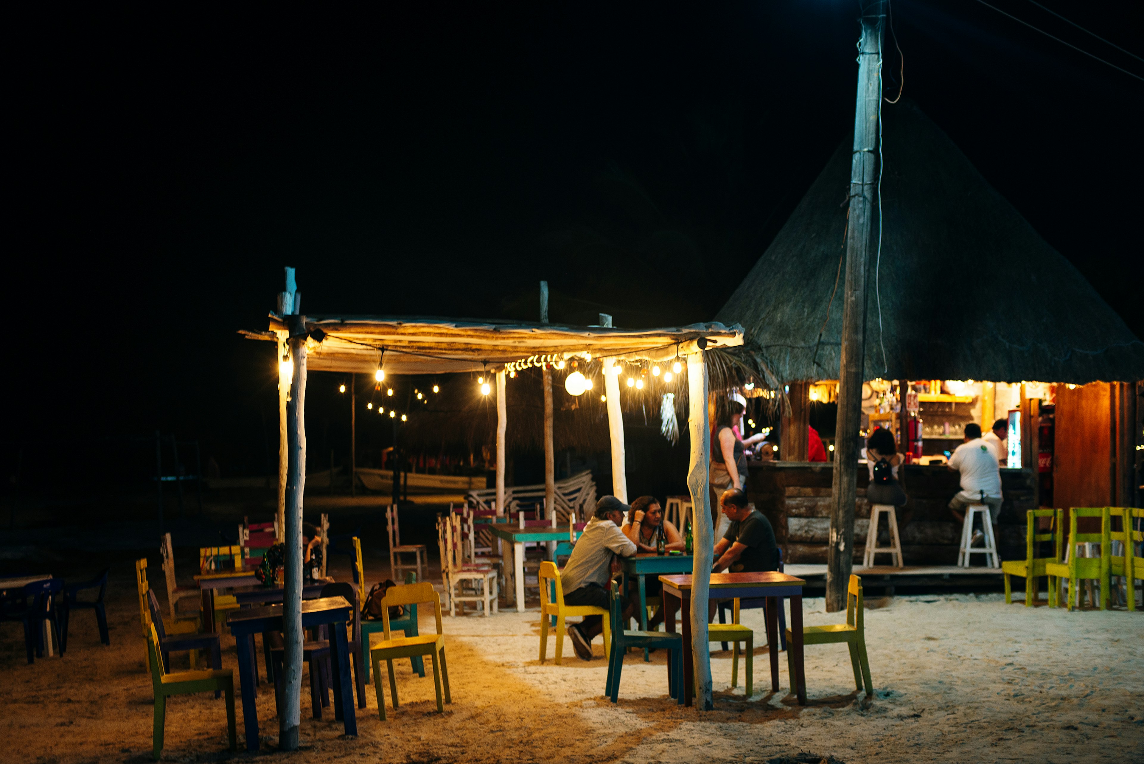 People sit at outdoor tables by night a bar on the beach in a tropical destination