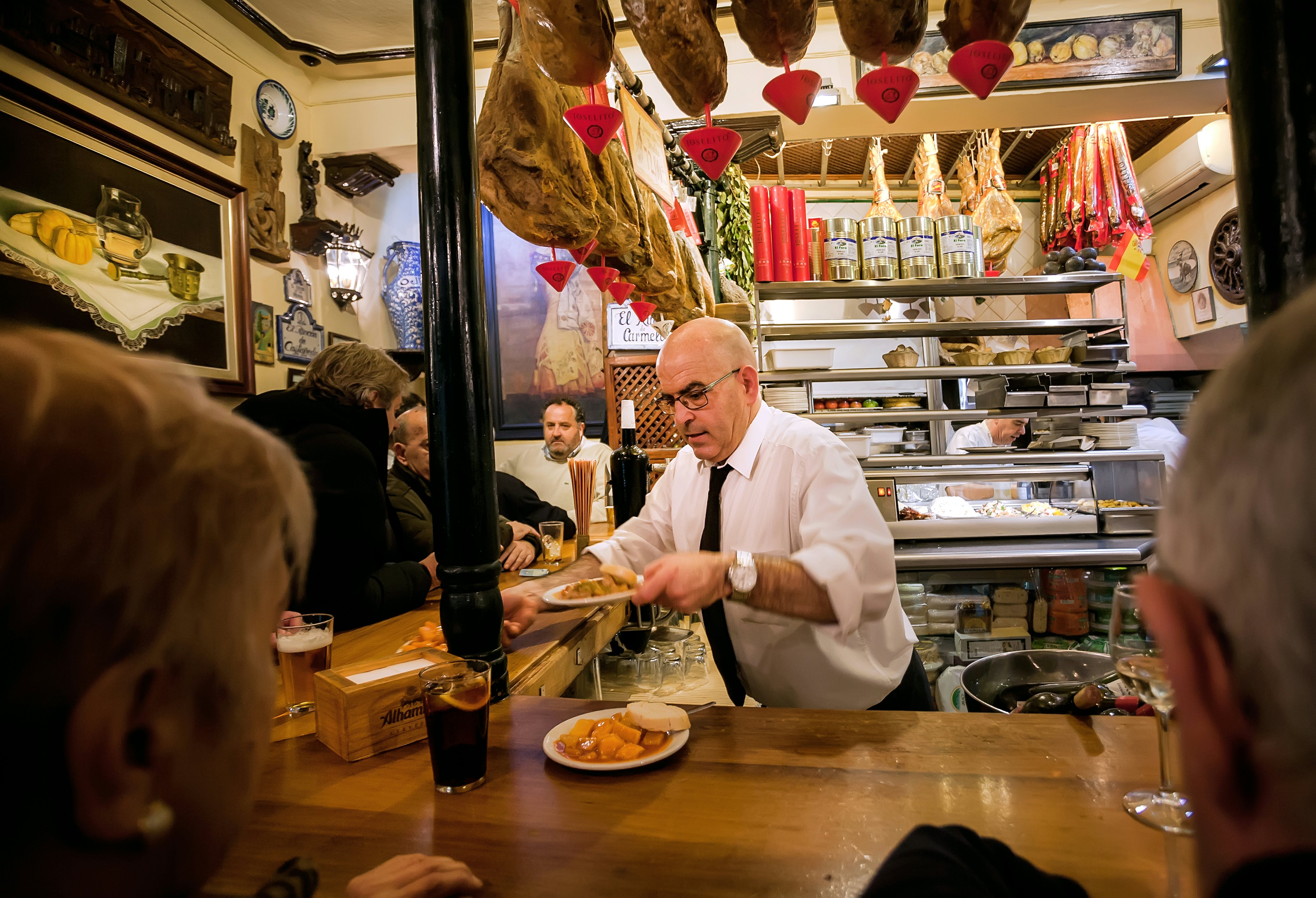 A waiter serves tapas and vermouth to diners at the bar in a rustic Spanish restaurant.