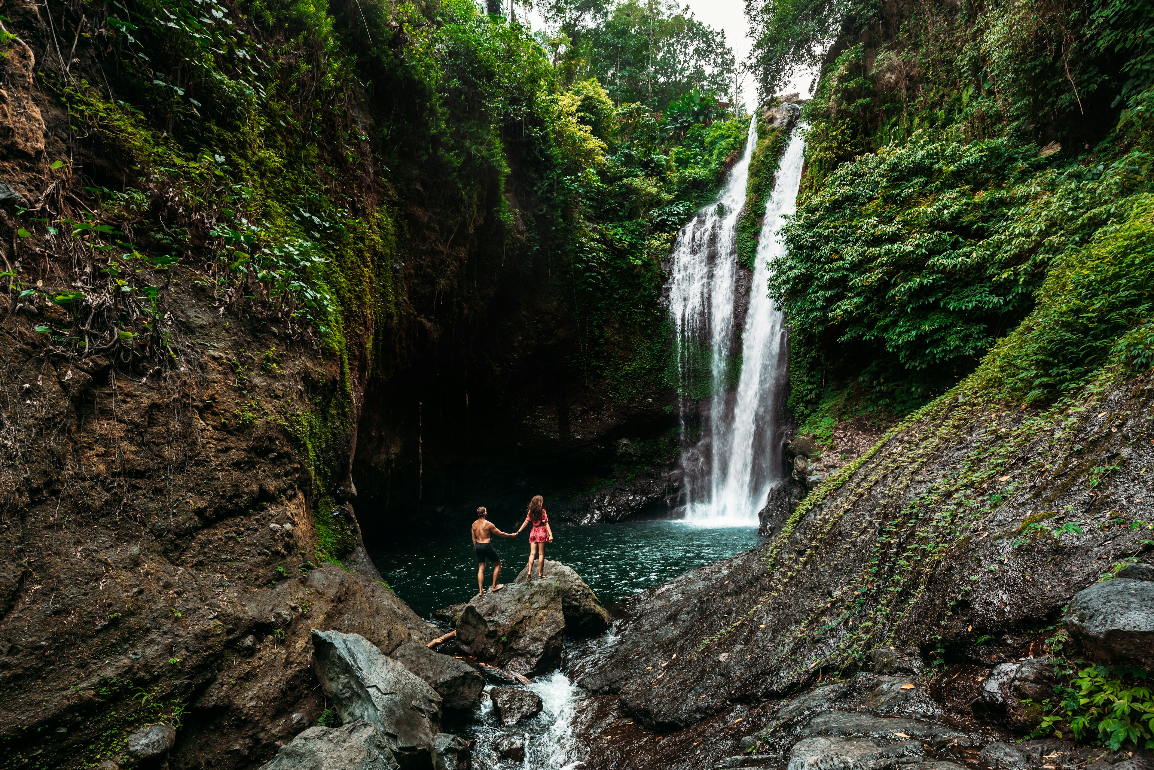 A wide shot of a couple holding hands as they gaze at a tall waterfall pouring into a pool surrounded by rocks from the green jungle above