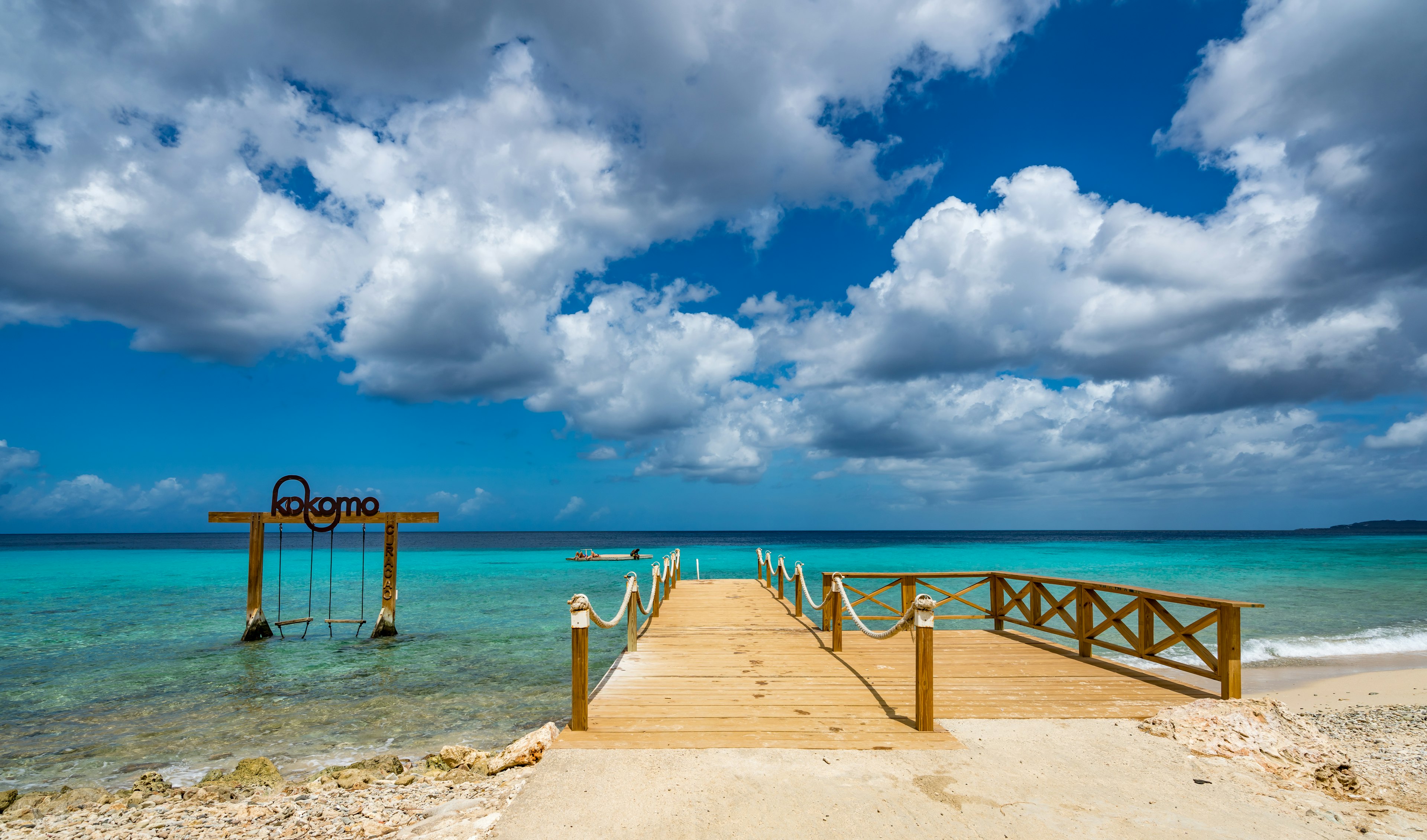 A small wooden pier leads out to a turquoise ocean next to two swing seats set in the sea