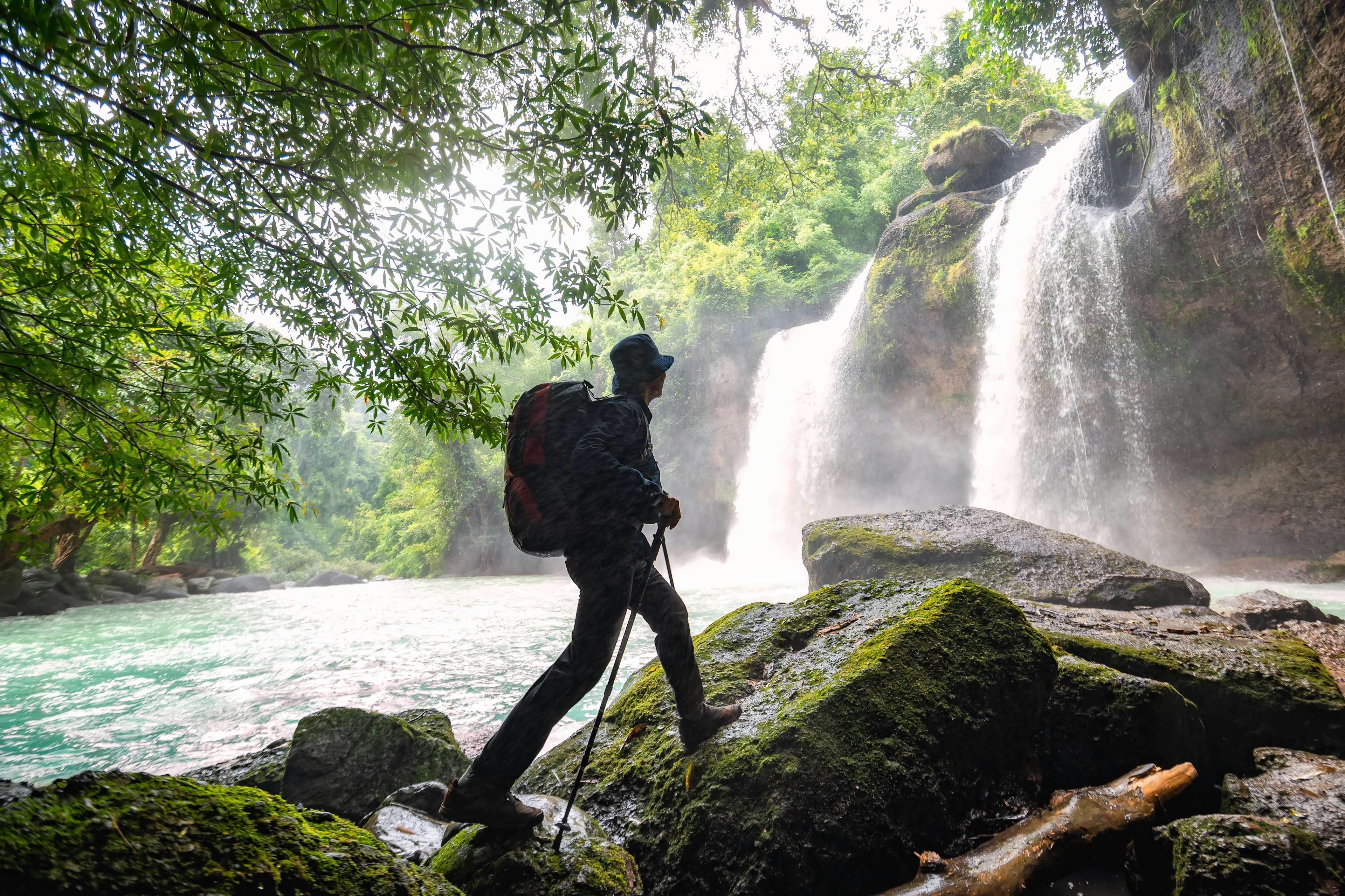 A hiker with poles and a large backpack is pictured in silhouette as he walks on mossy rocks in front of a tall waterfall in the rainforest