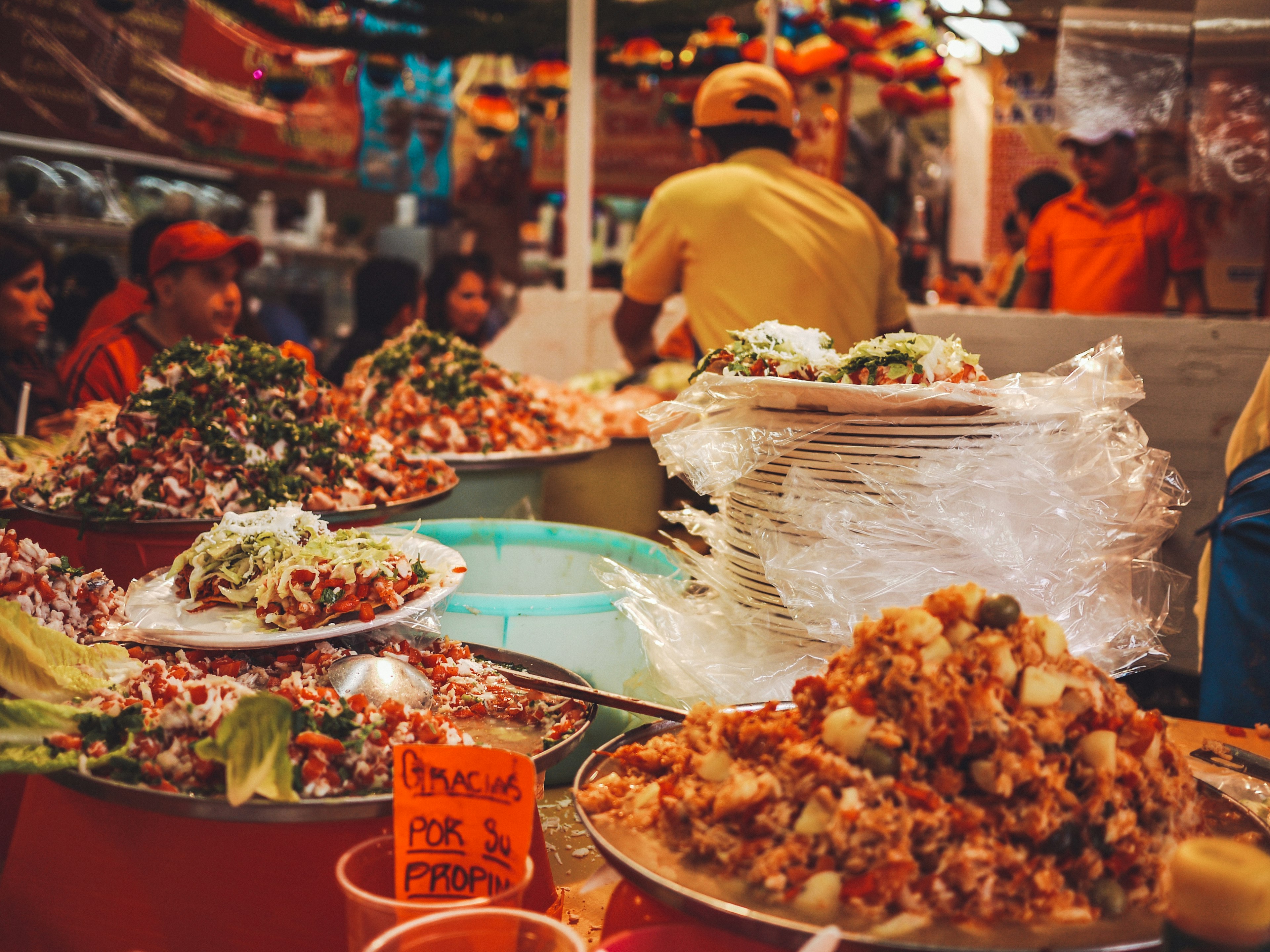 Tostadas and other freshly prepared foods for sale at a market stall, with a chef and customers visible in the background