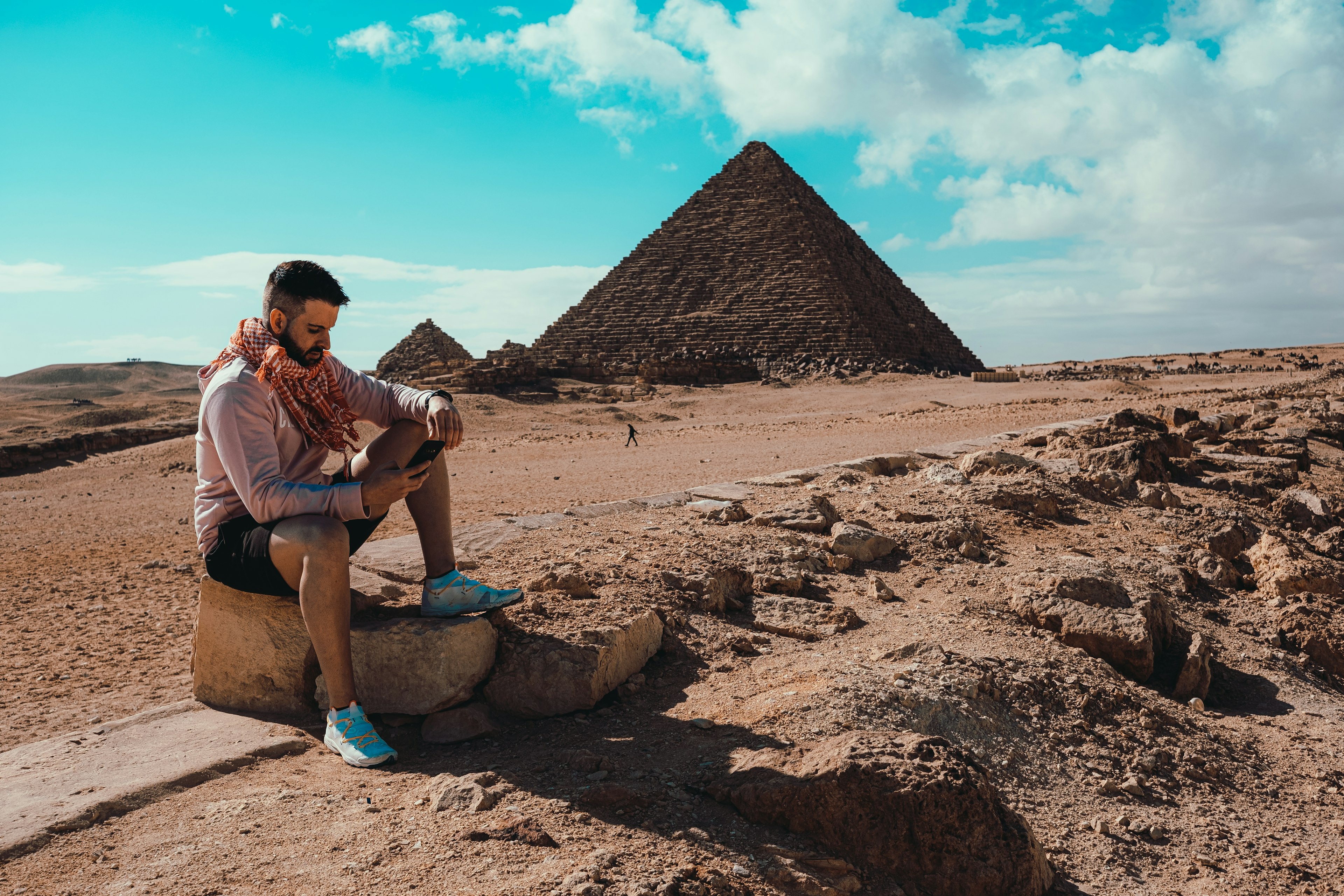 A man, sitting on a rock in front of a huge stone pyramid in a desert-like landscape, looks at his phone