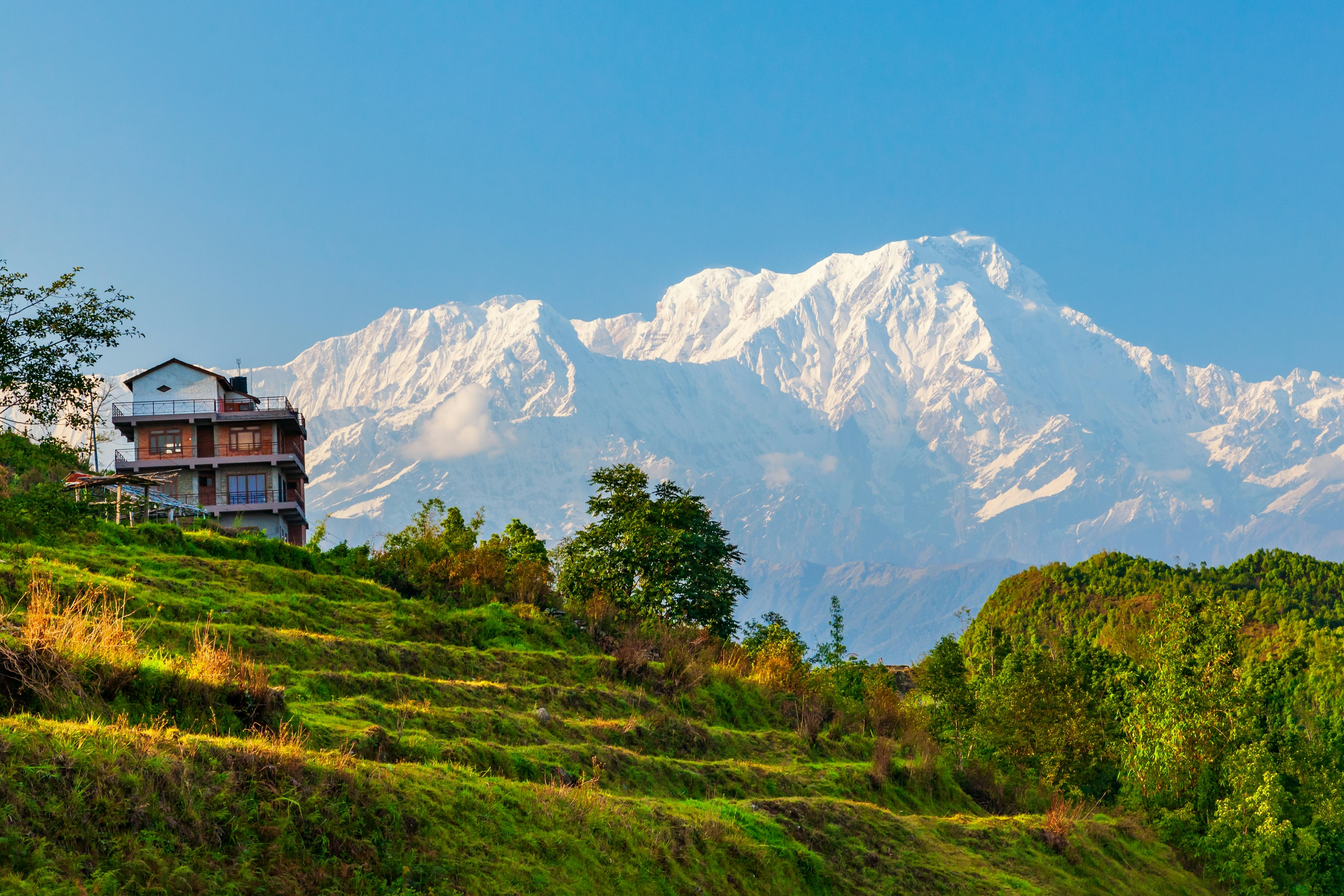 A grassy hill leads up to a tall house, backed by snow-capped mountains against a blue sky