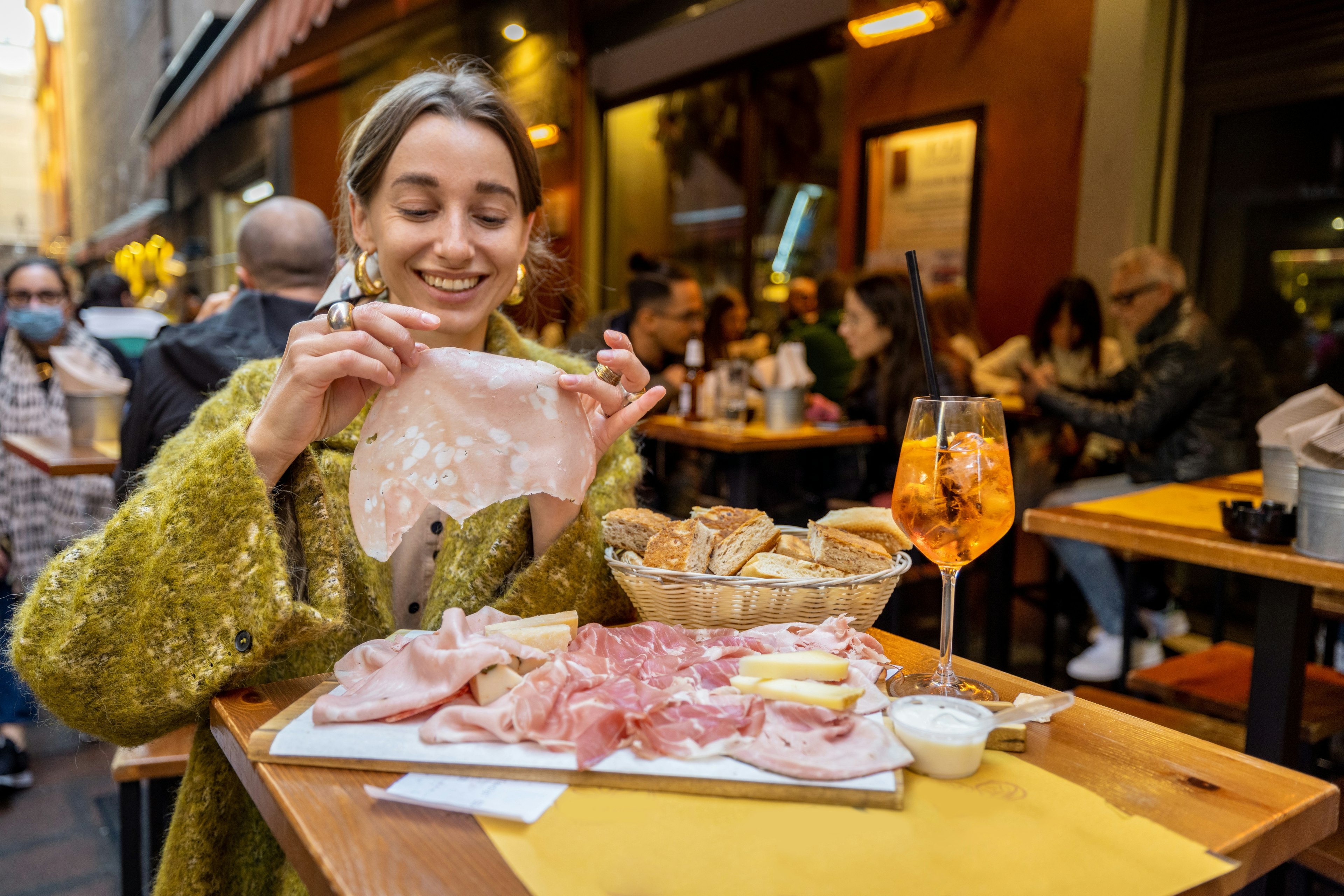 Woman having fun, while eating meat plate at outdoor bar in Bologna city. Girl holds mortadella, famous italian sausage.