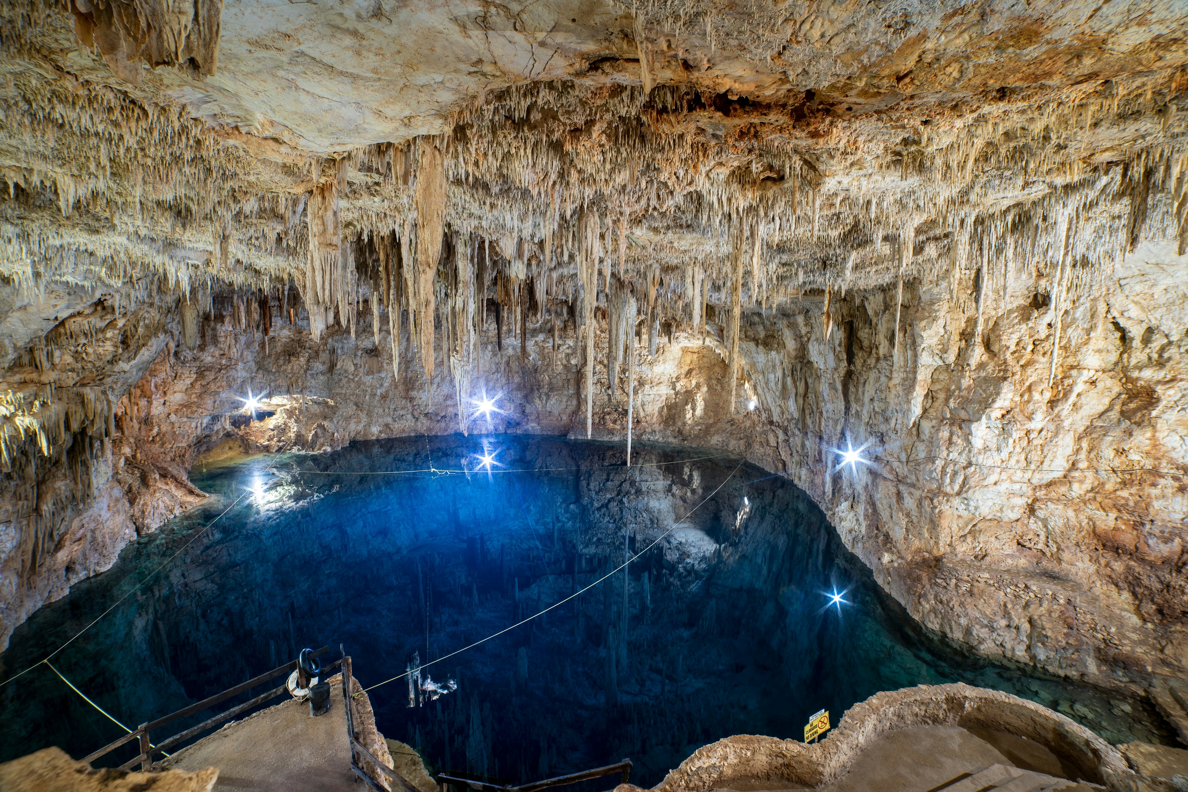 A cenote (underground pool) is illuminated, with spotlights making the water look deep blue. Countless stalactites hang from the ceiling of the cavern.