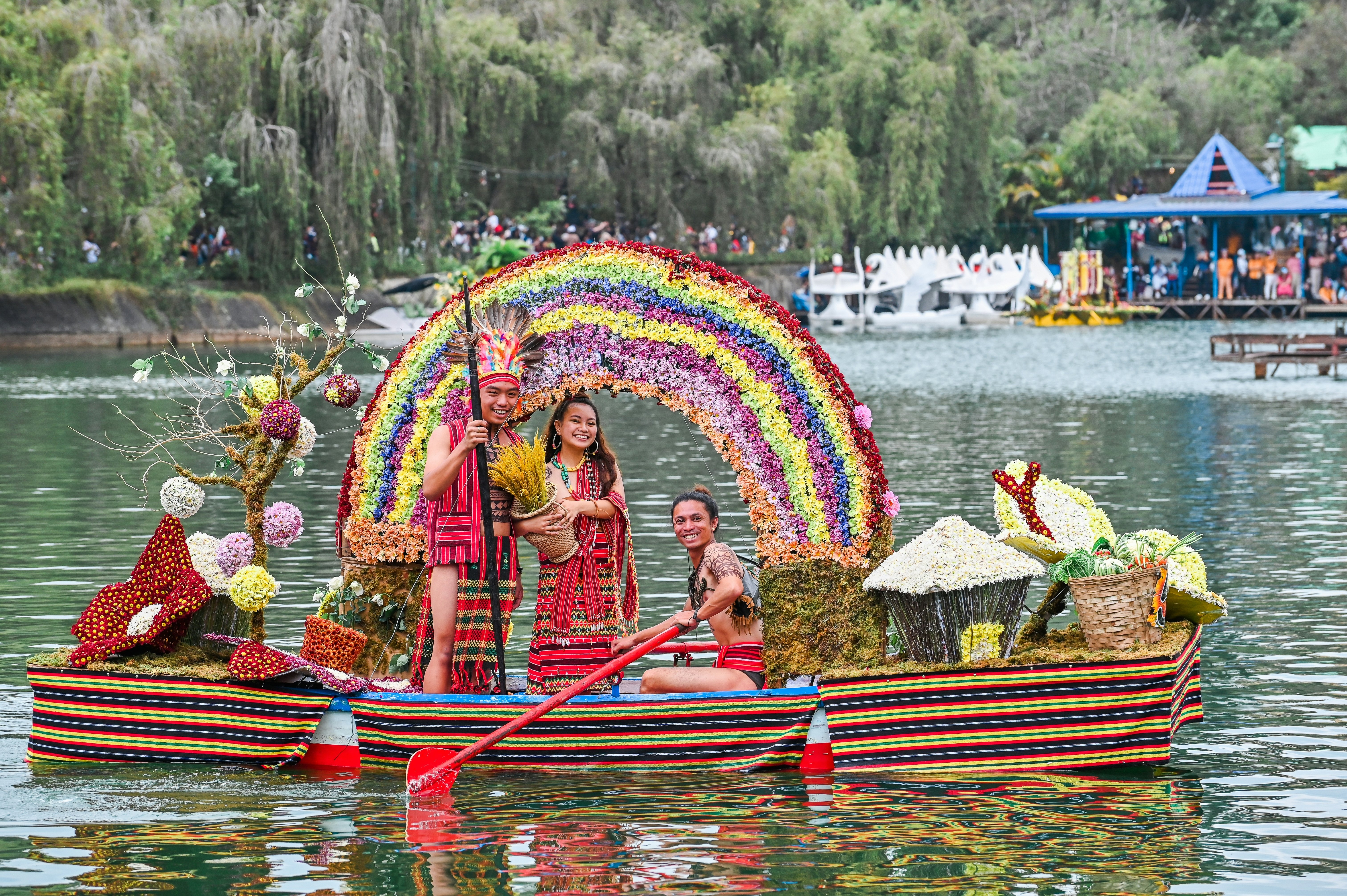 People in traditional costumes paddle a boat covered in displays of flowers on a lake during a festival