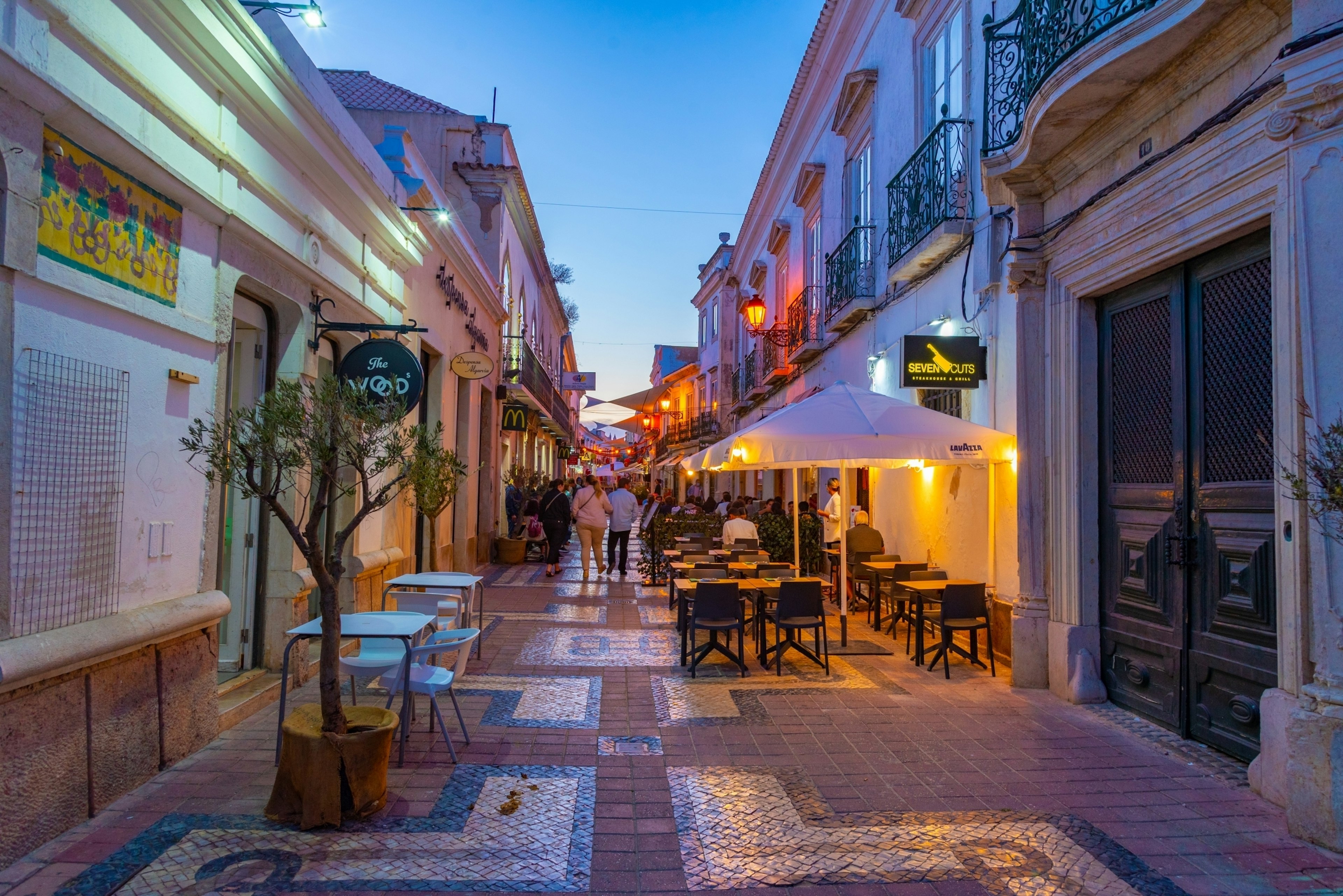 People walk down a tiled, pedestrianized street in the evening past outdoor tables and other bars and restaurants