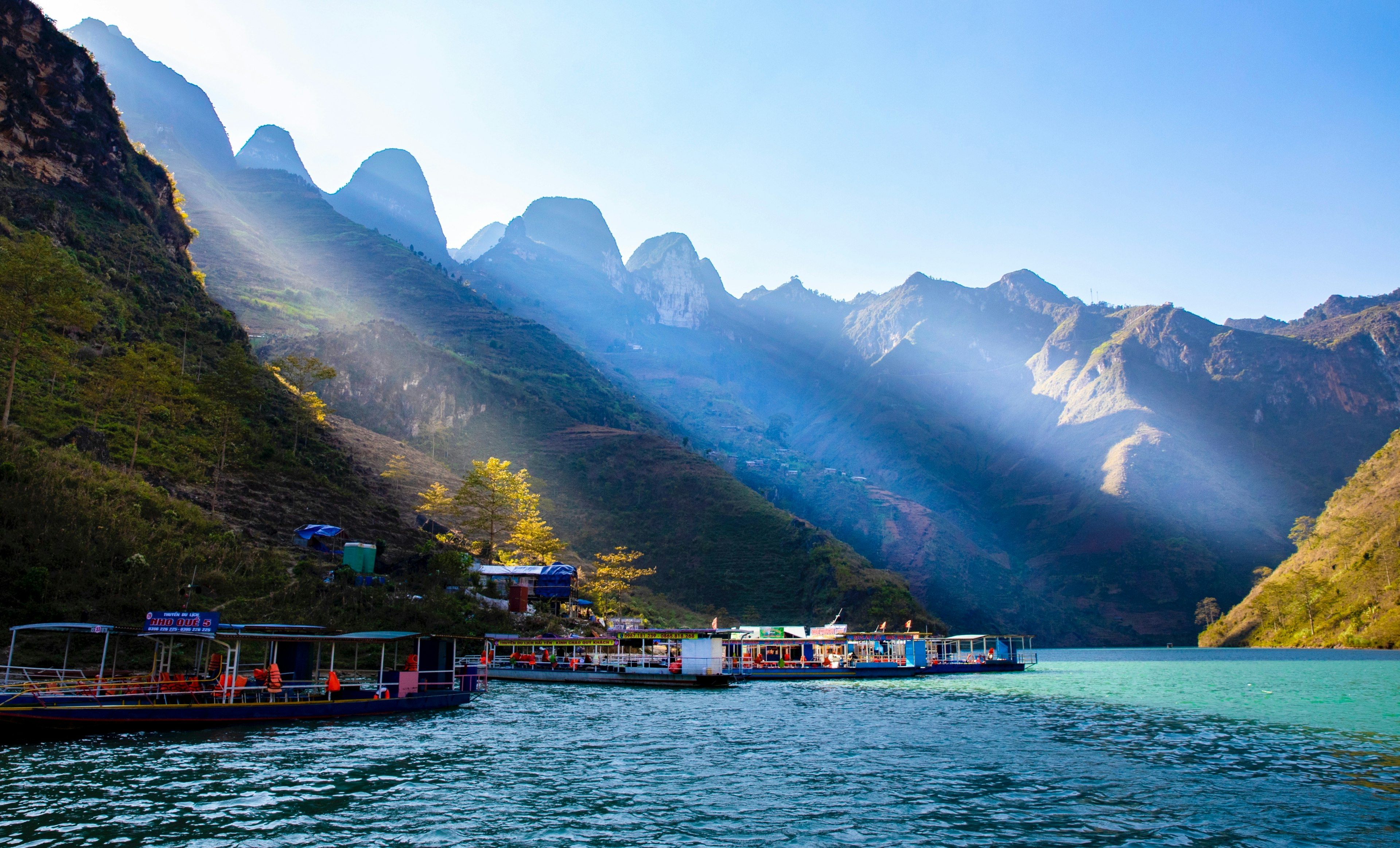 A view of passenger boats moored on a riverbank with spectacular karst mountains in the distance, all illuminated by rays of sunlight