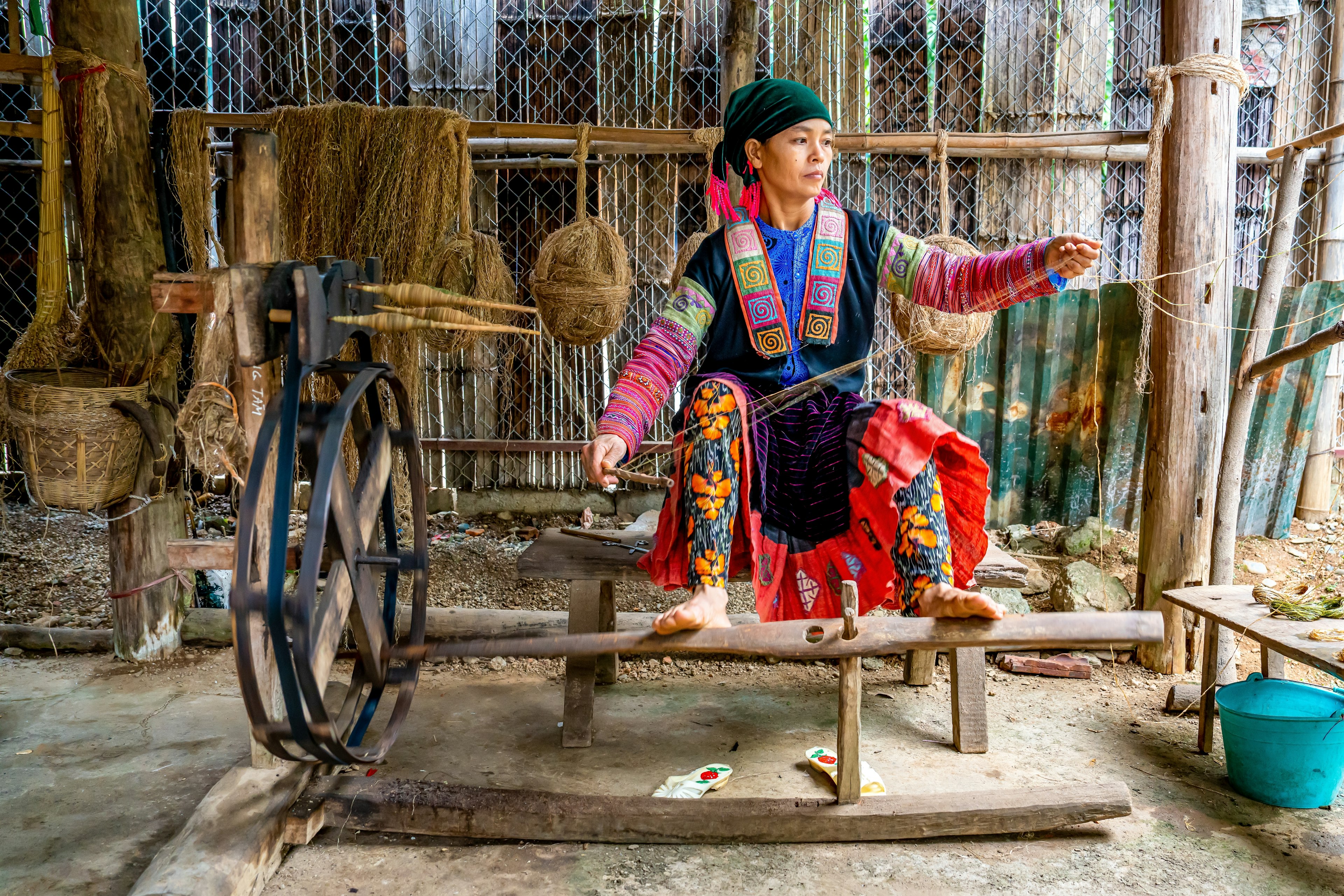 A Hmong woman in traditional, brightly colored dress weaves at a loom in a wooden building