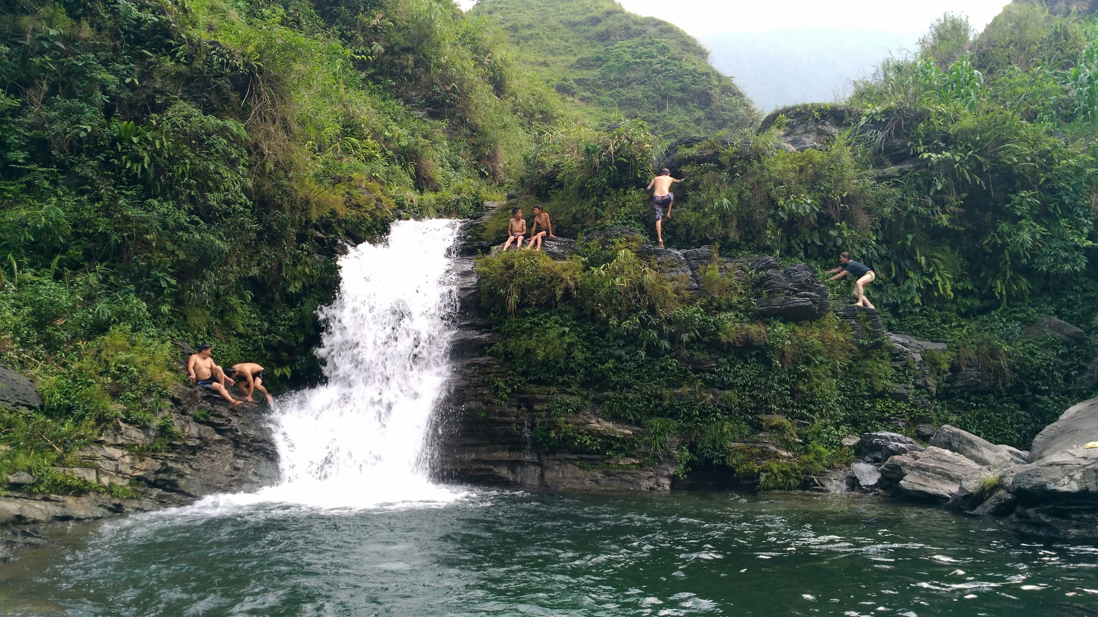 People clamber along rocks next to a large waterfall and swimming hole in the mountains. Greenery covers the landscape.