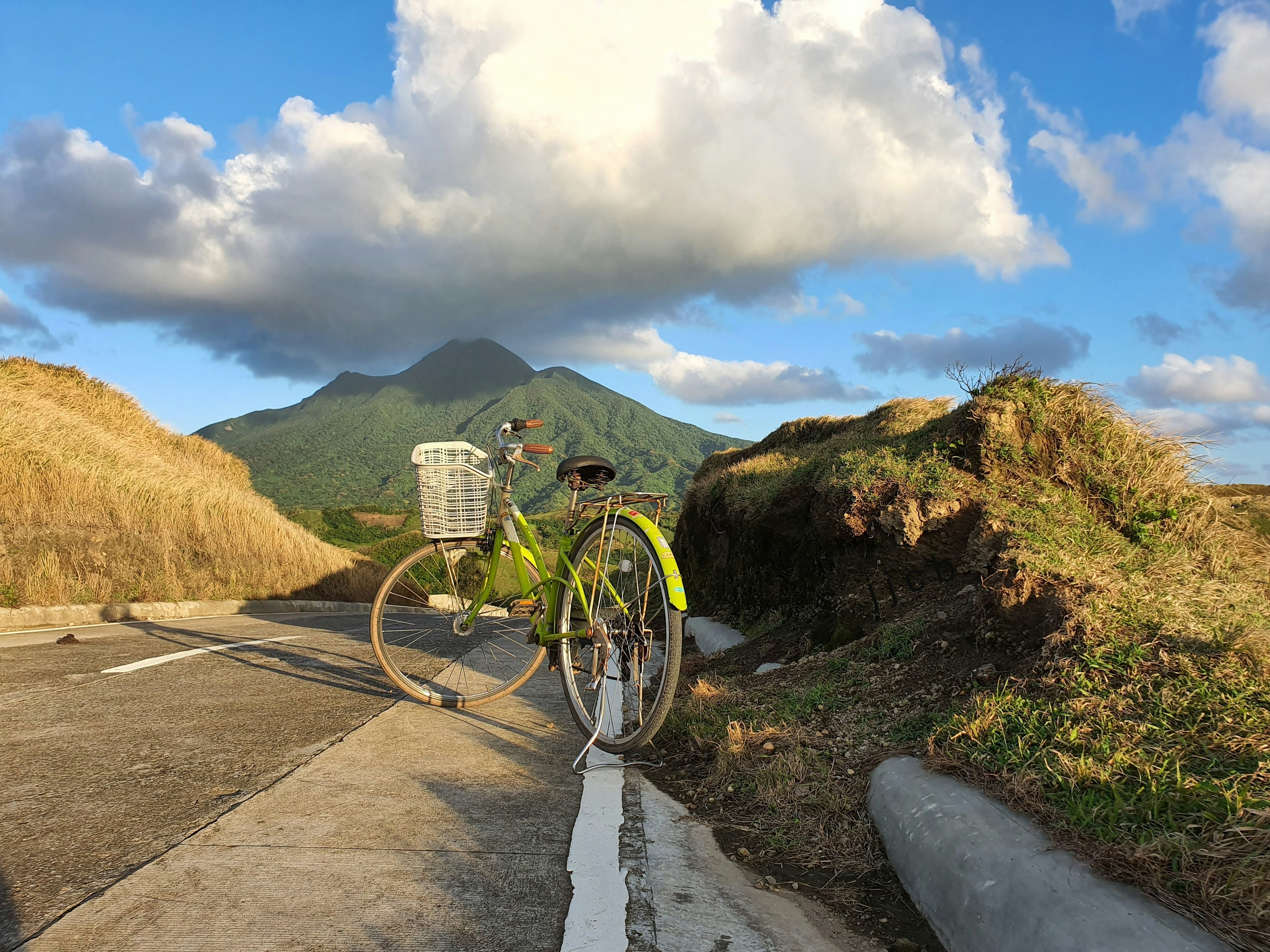 A bicycle with a front basket is on the side of a mountain road, with a mountain peak under cloud cover in the distance