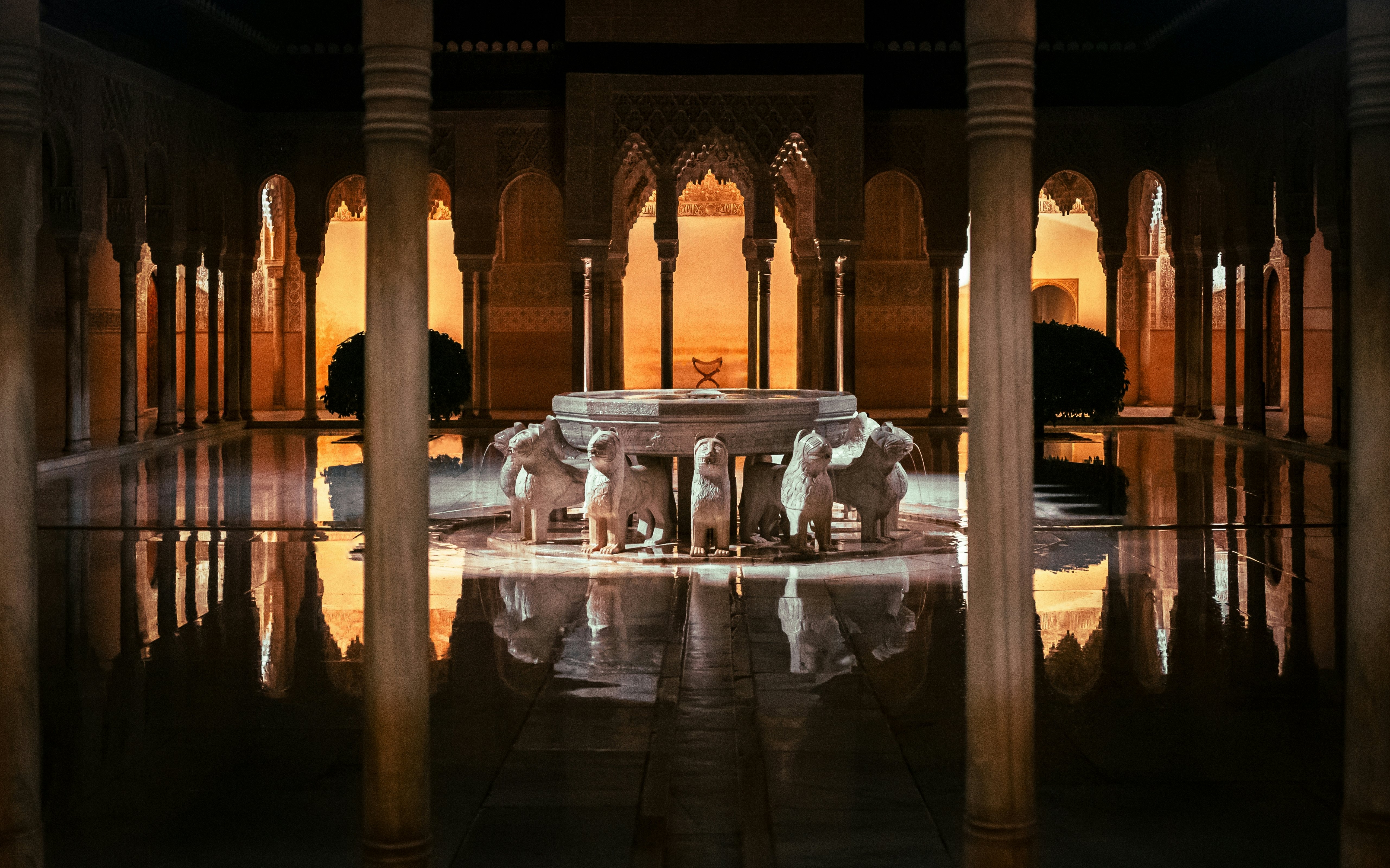 A fountain encircled by sculptures of lions is in the center of a courtyard; the stone is wet, and the fountain and surrounding columns and archways are reflected on the water's surface.