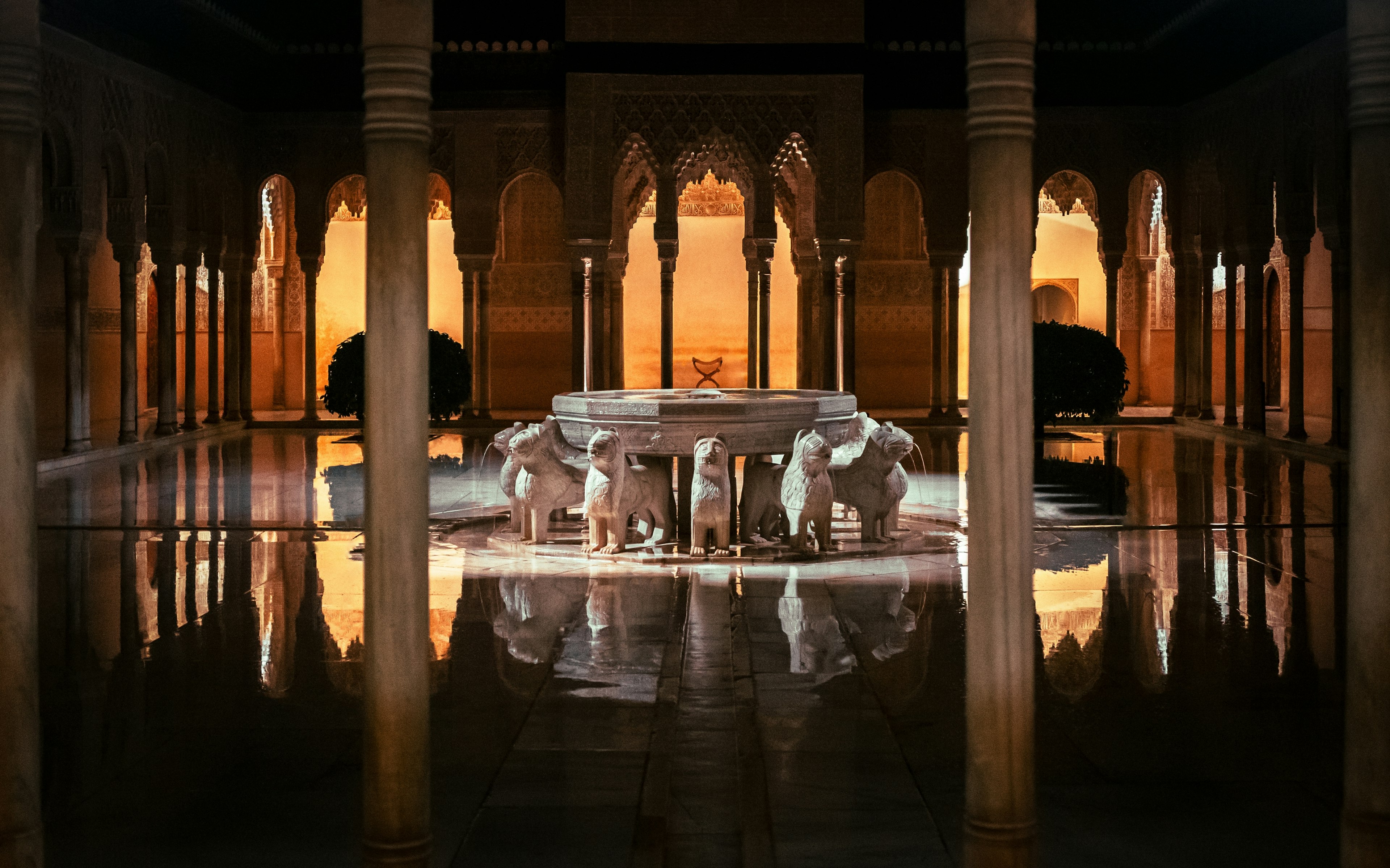 A fountain supported by carved lions in a shallow pond surrounded by slender columns and arches, illuminated dramatically at night