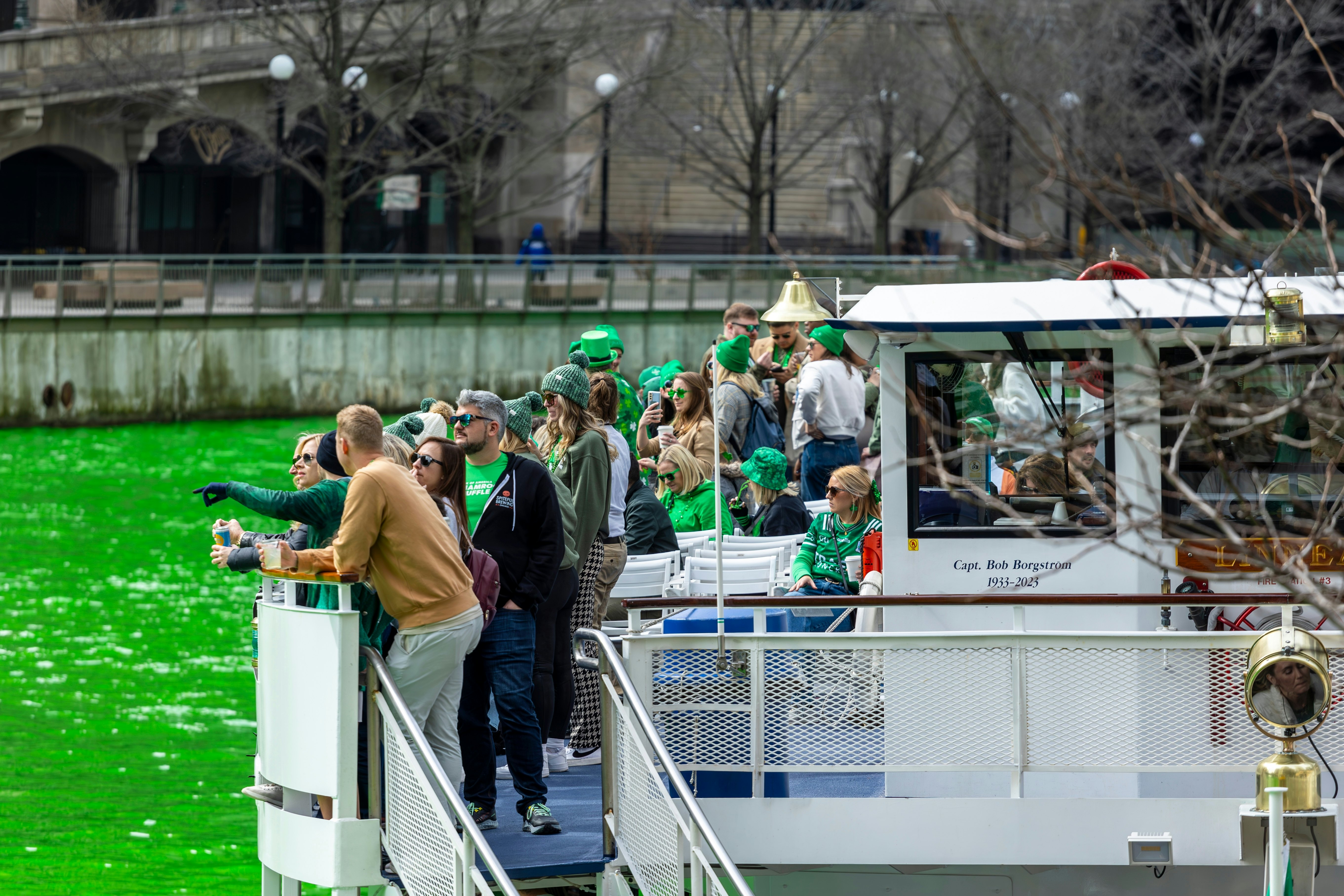 People dressed with bright green accessories stand on the deck of a boat on a river that's been dyed green