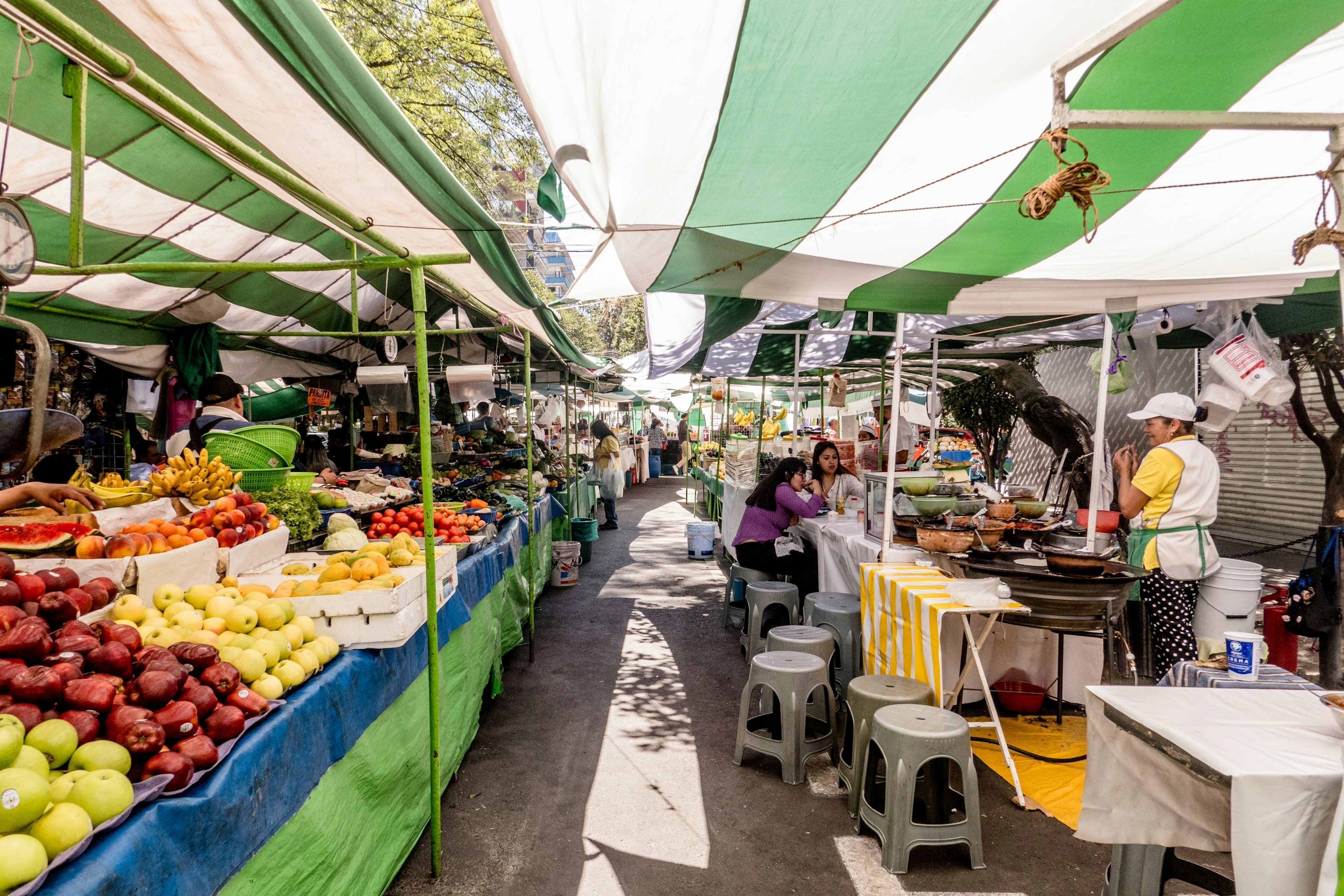 An open-air tianguis market features stalls selling fresh fruits and vegetables, and vendors selling fresh food and a seating area with small stools