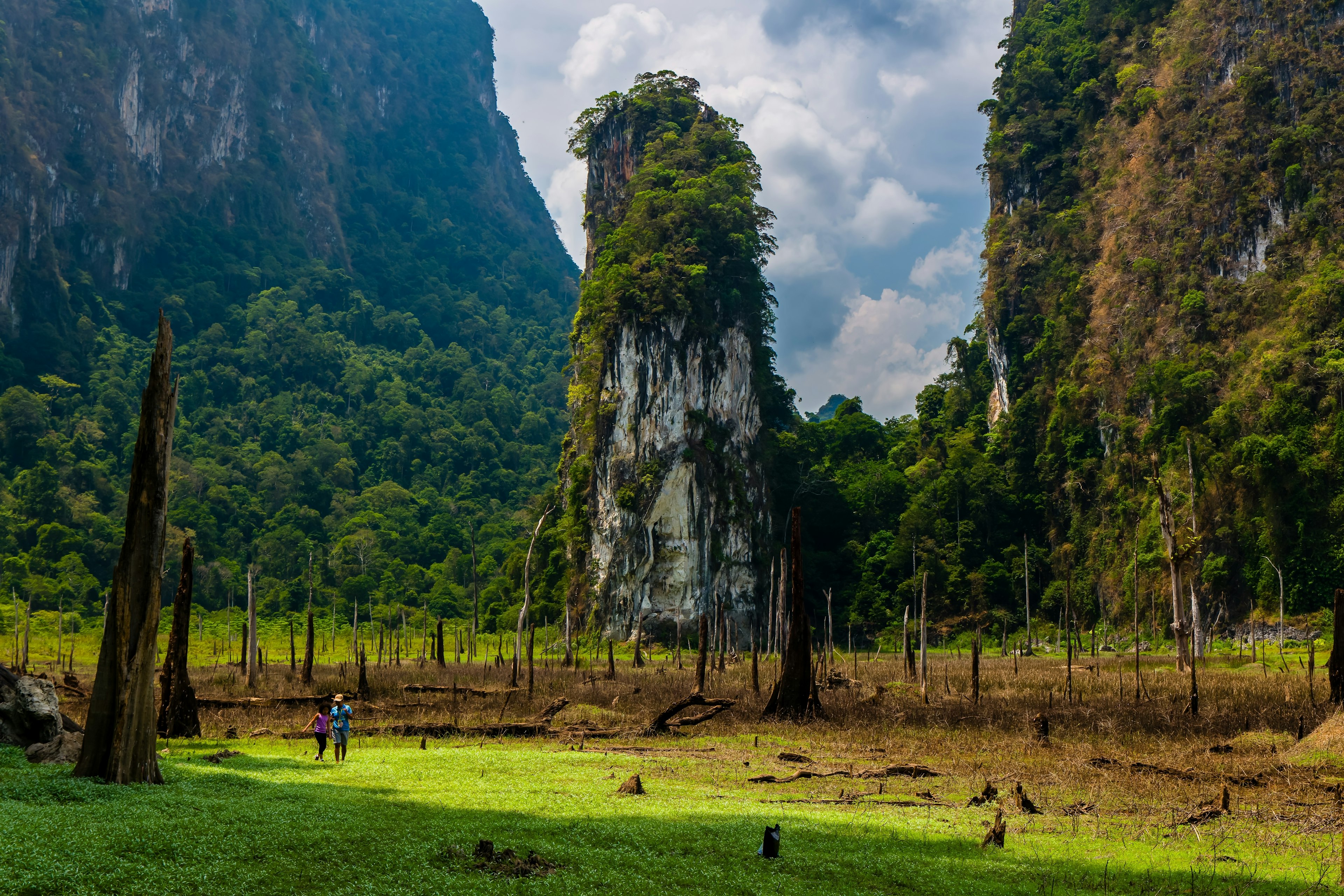 A wide shot of two hikers crossing a grassy area in front of a tall karst column, with larger, vegetation-covered rocky mountains rising on either side