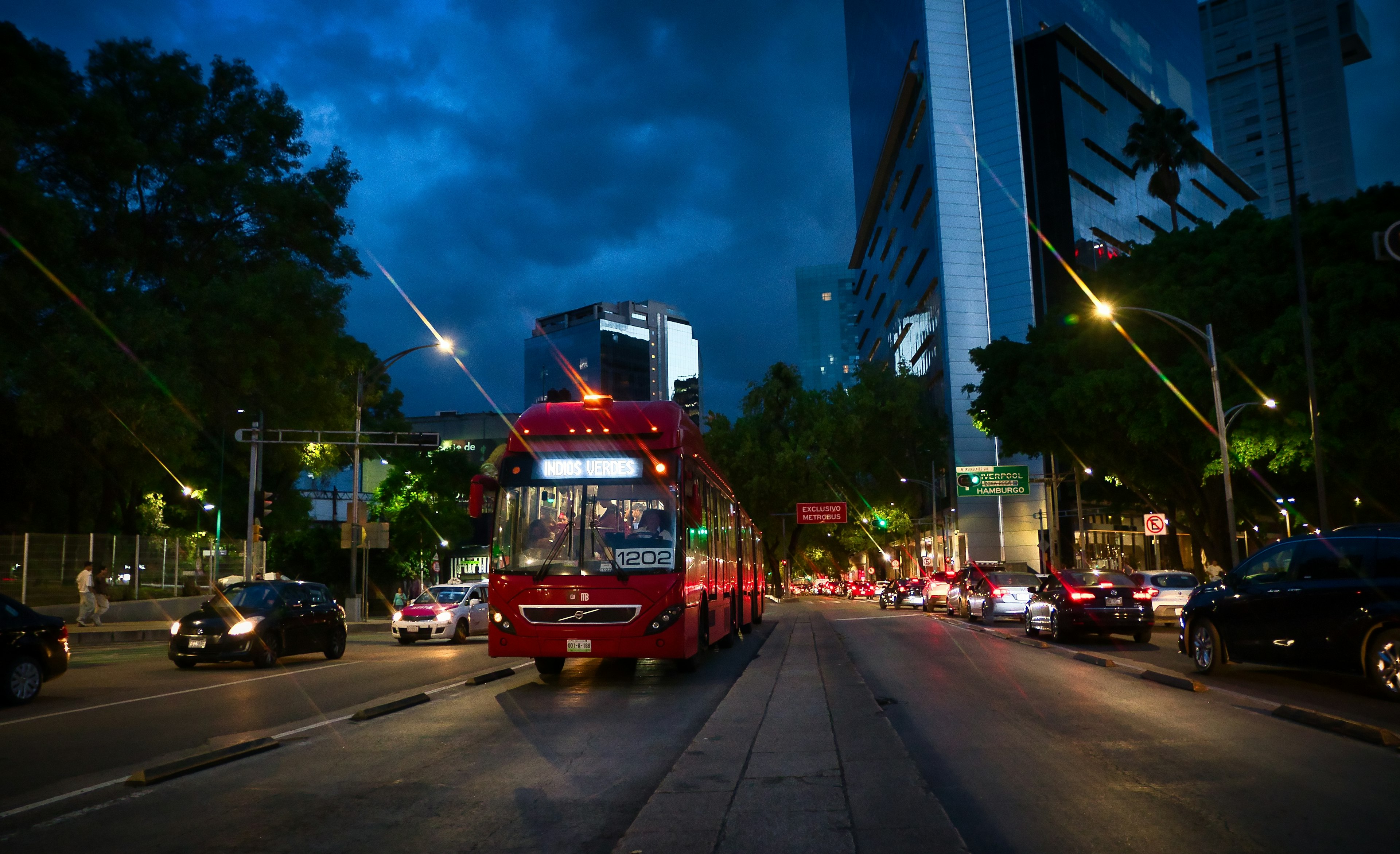 A red city bus drives down a wide boulevard at night, with other cars and skyscrapers visible