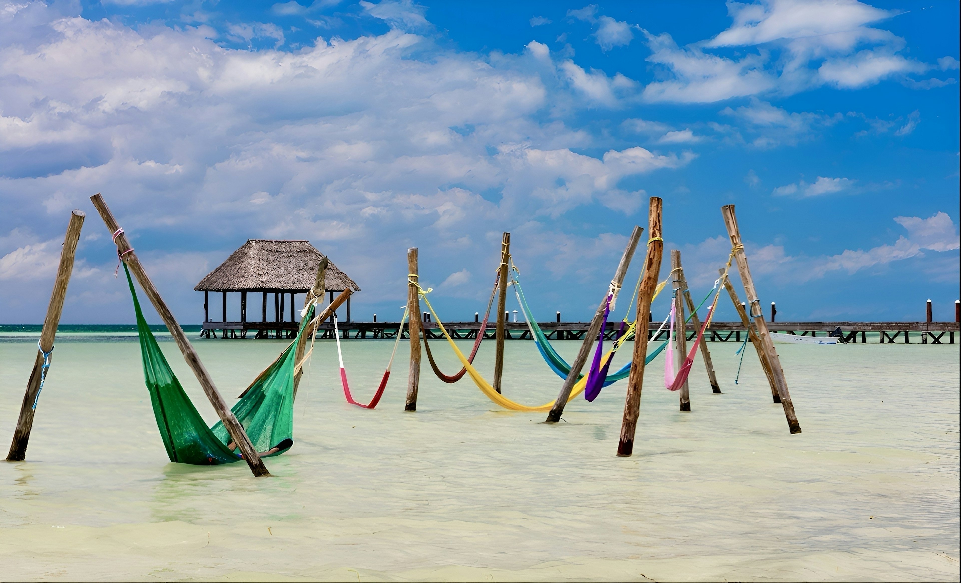 Colorful hammocks hang from poles in the shallow water of a tropical beach. A pier with a pavilion is visible in the background of the image.