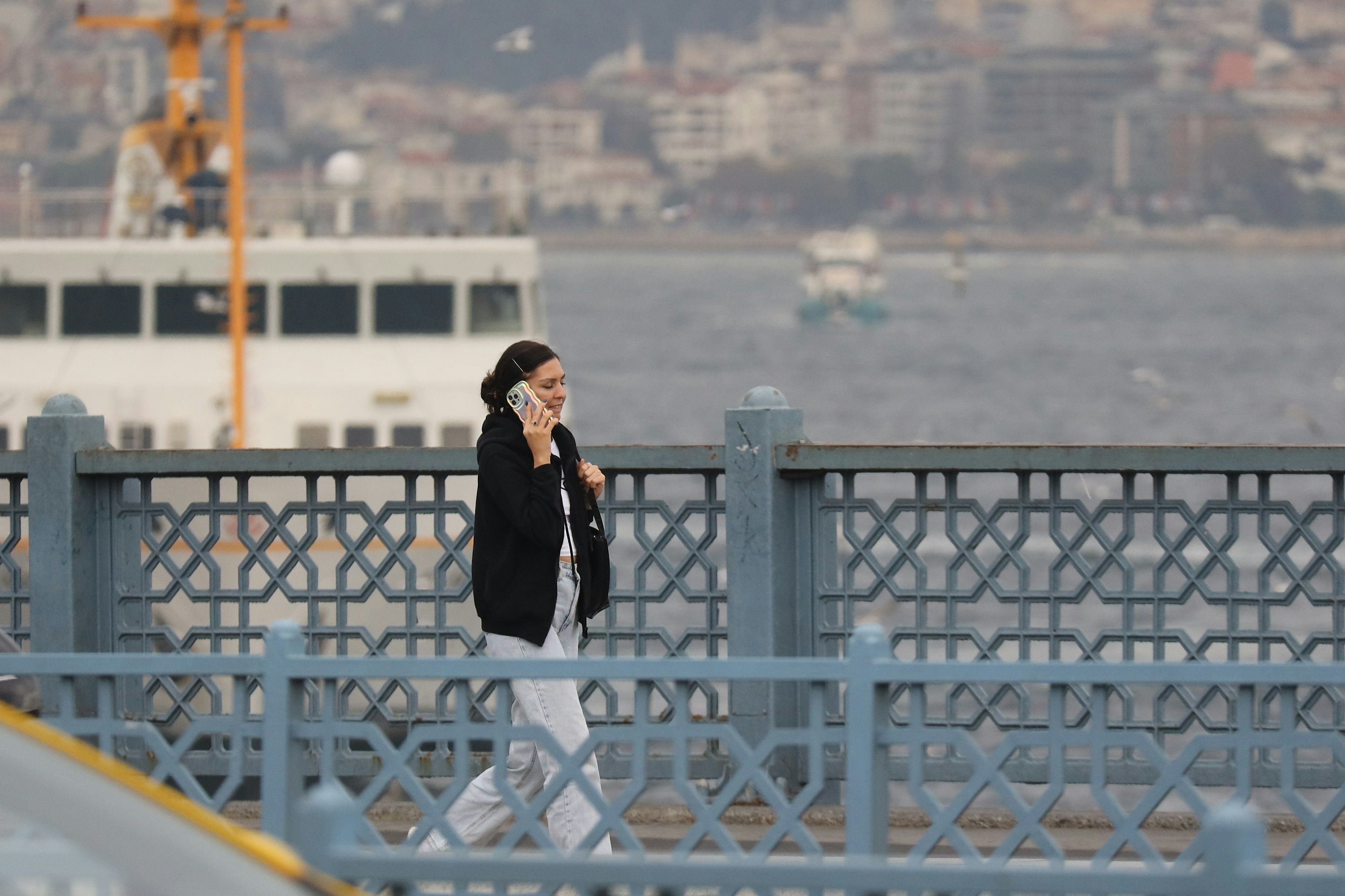 A woman talks on her phone while walking on a bridge with a ferry in the background,