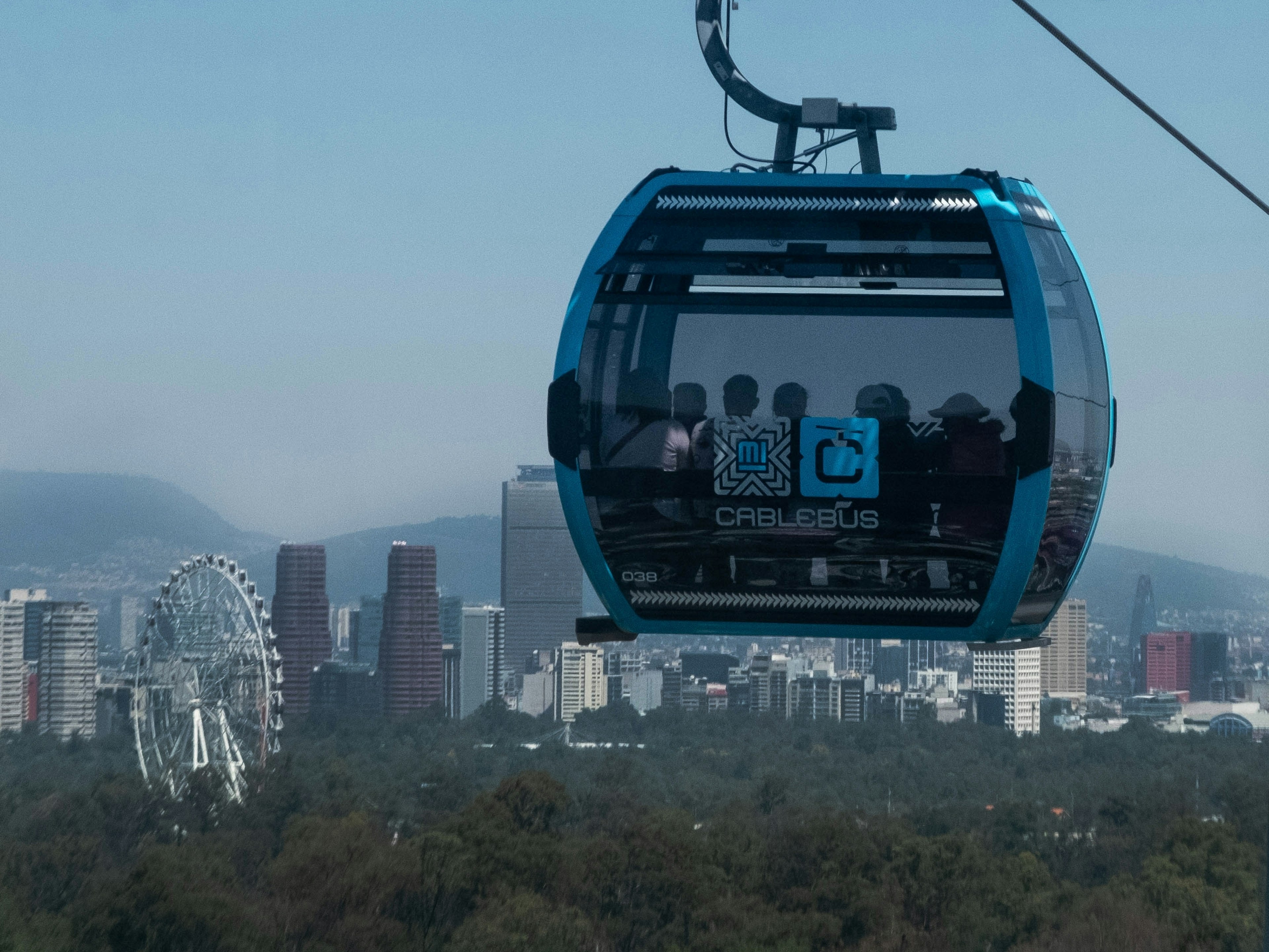 Passengers in a cable-car cabin look out over a city skyline, with a Ferris wheel, skyscrapers and distant mountains visible