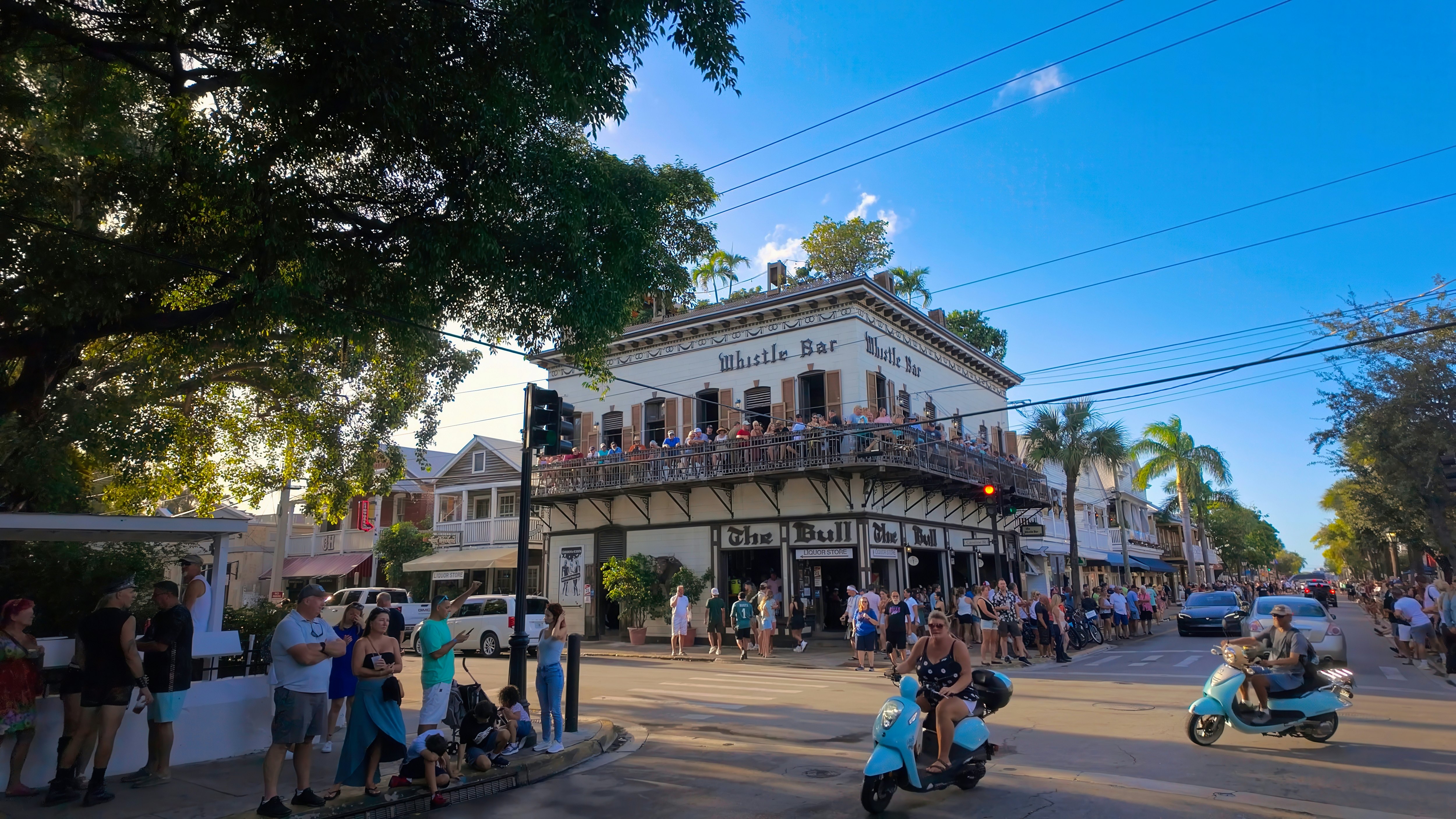 People on foot and on scooters walk by an intersection and a building that houses a bar with a wraparound balcony. Palms and other trees line the side streets.