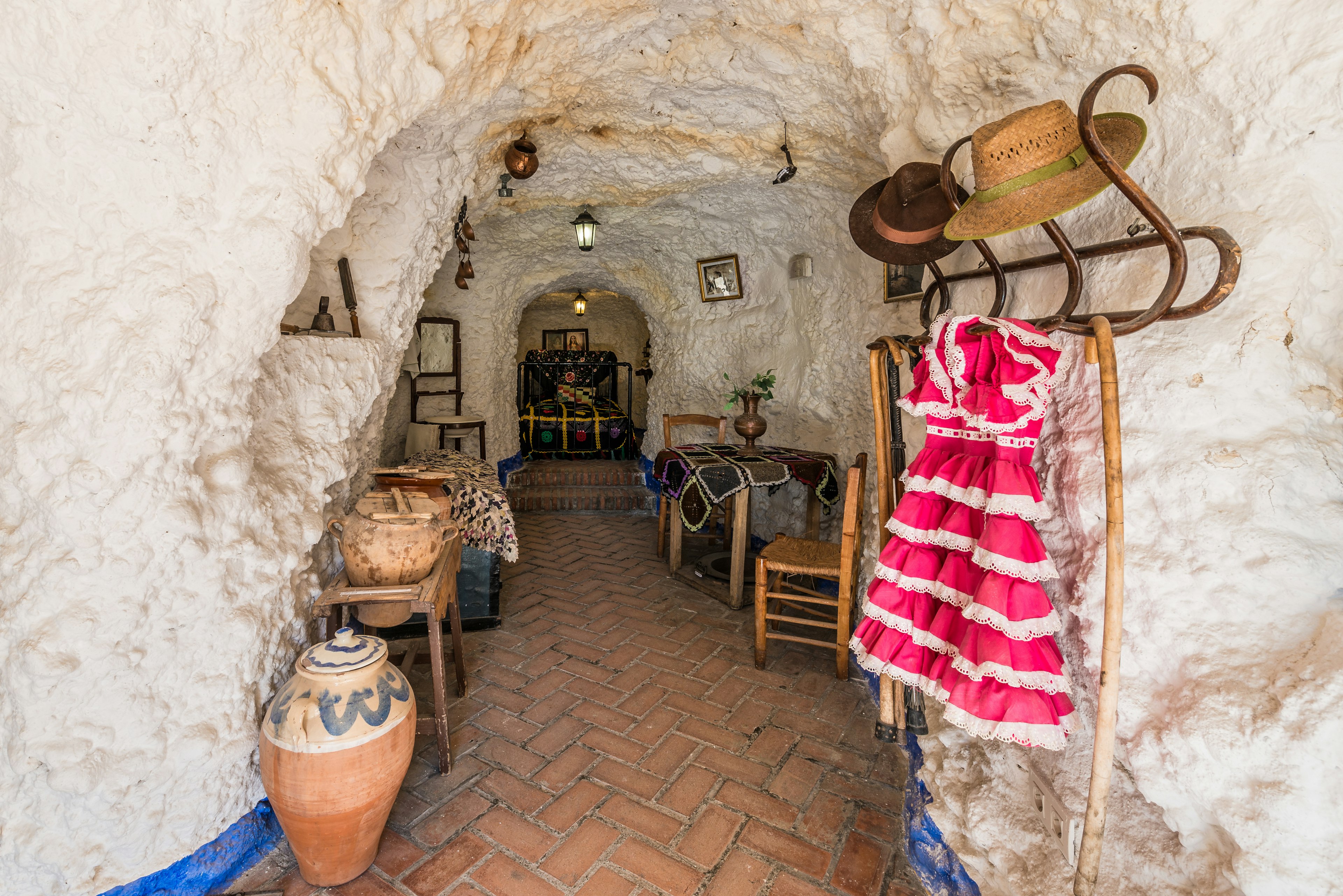 A narrow living space is carved into a rock cave. Earthenware jugs, a pink dress, table, chairs and other furniture and furnishings are visible in the small space.