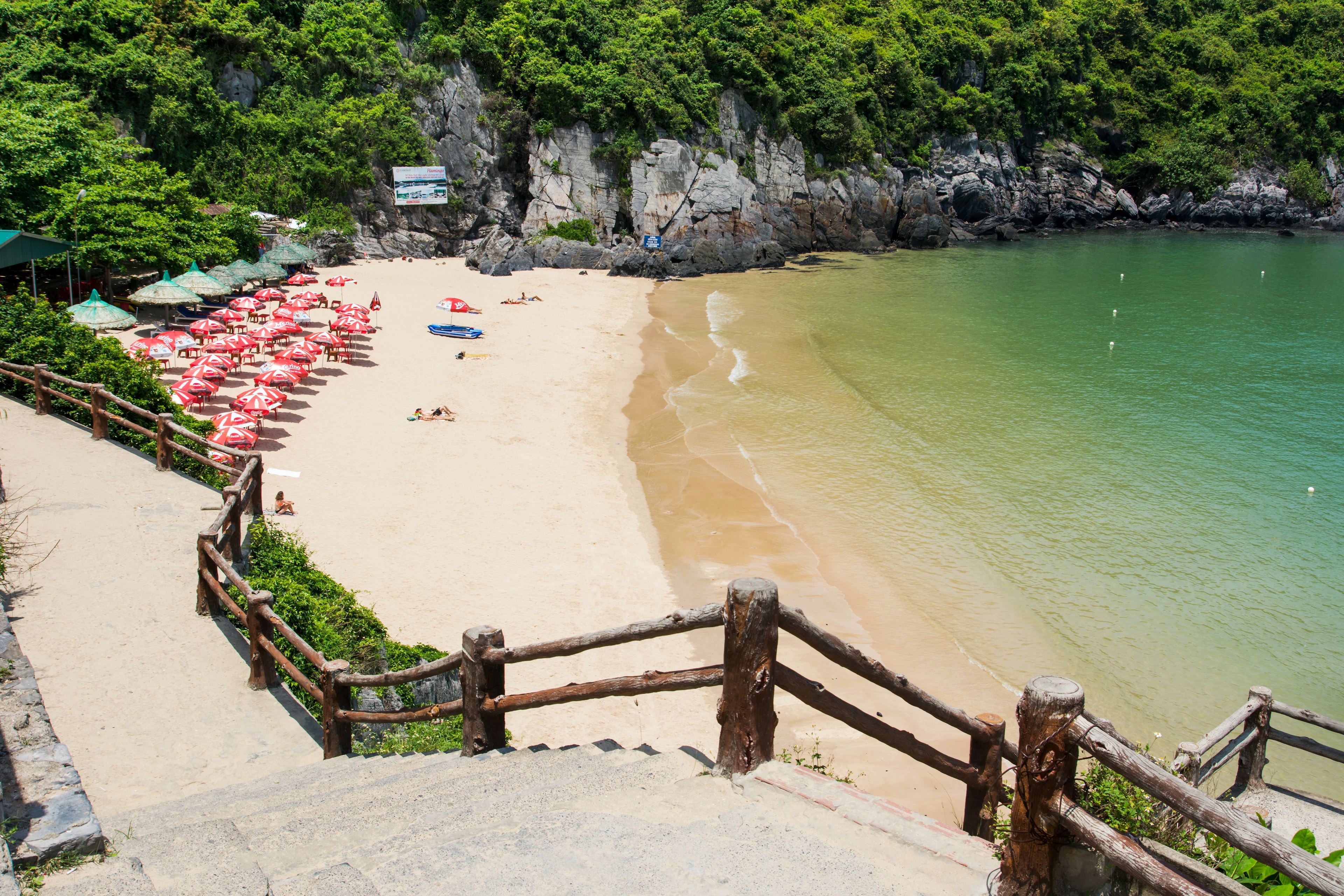 A view from the top of a flight of stairs leading to a small beach with red sun umbrellas, on a cove lined with rock formations and green vegetation.