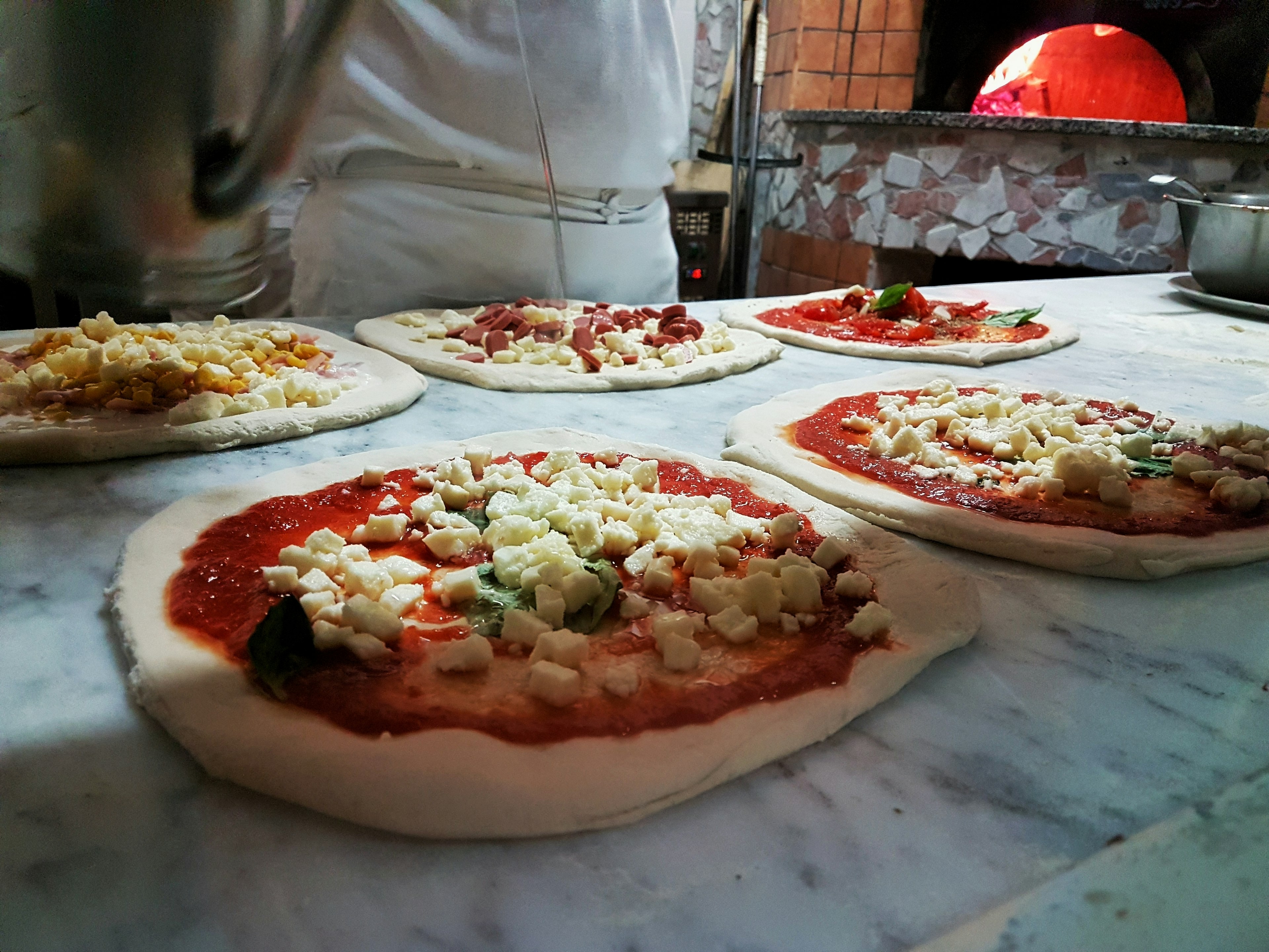 Five pizzas being prepared next to a wood-fired oven