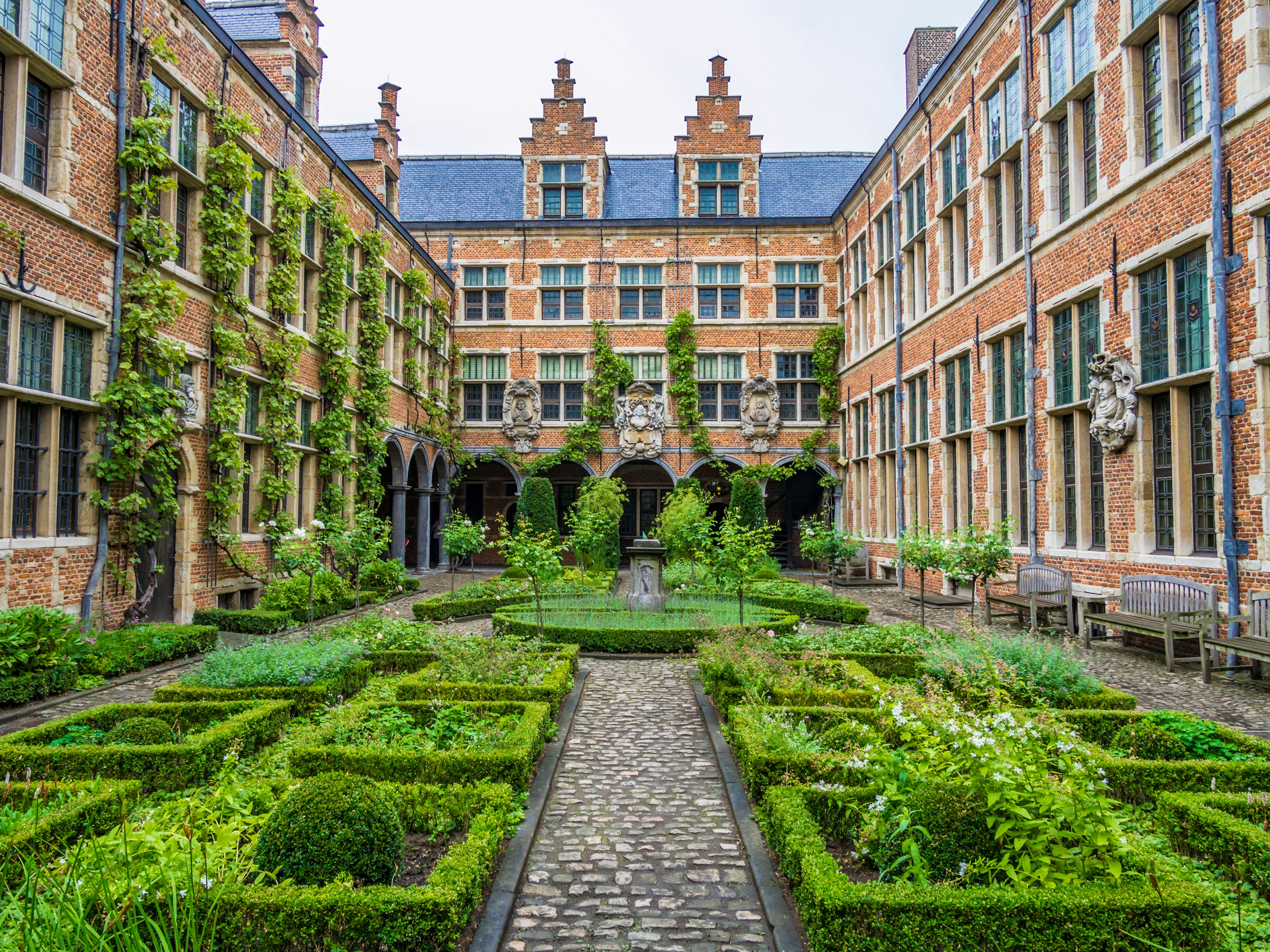 A courtyard of a medieval red-brick building with landscapes flower borders and a central fountain