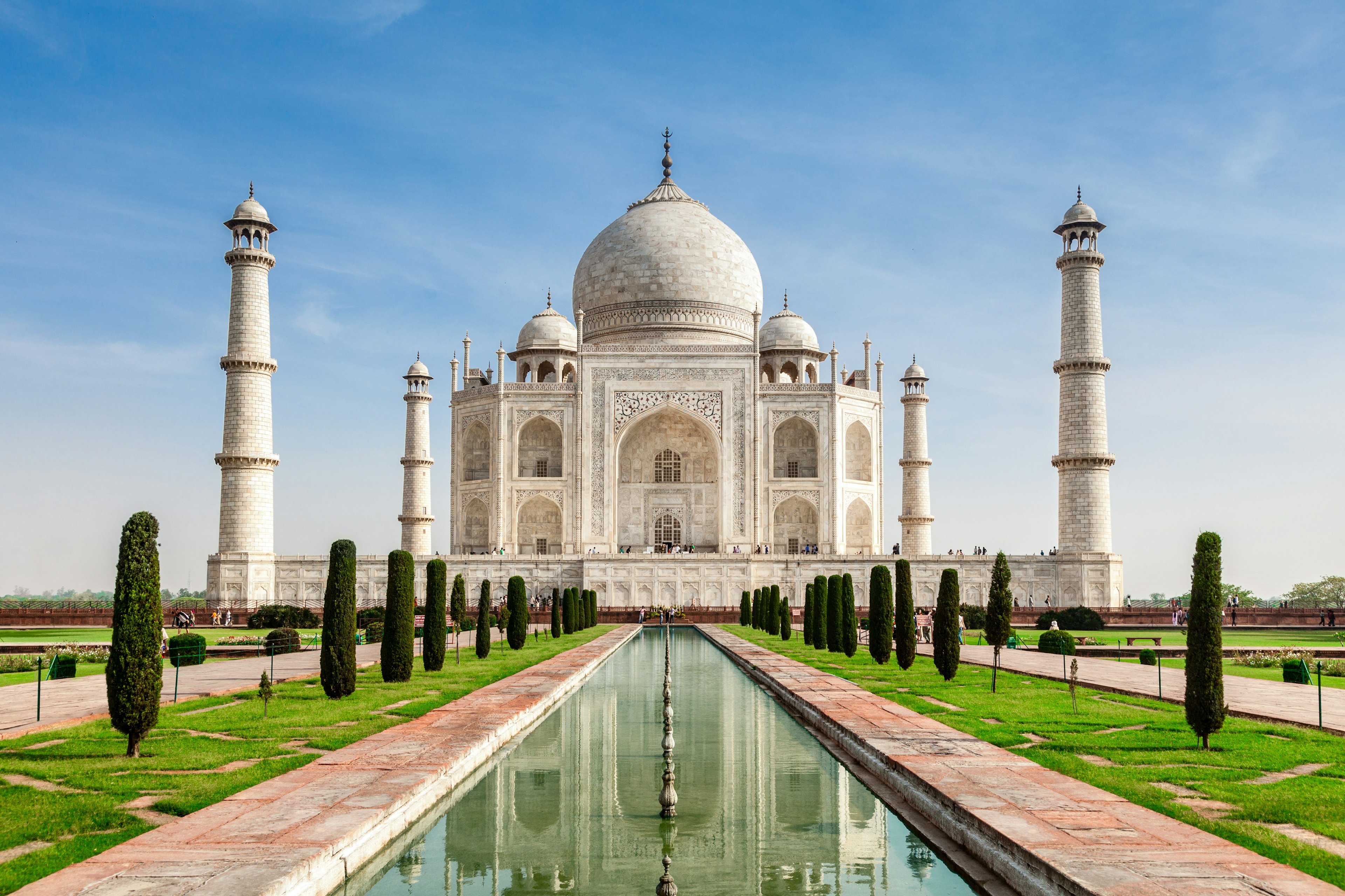 The Taj Mahal shot from the front in the middle of the day. The white marble structure, with its domed roof and four white pillars surrounding it, is partially reflected in the long, thin pond that runs towards the entrance of the building.