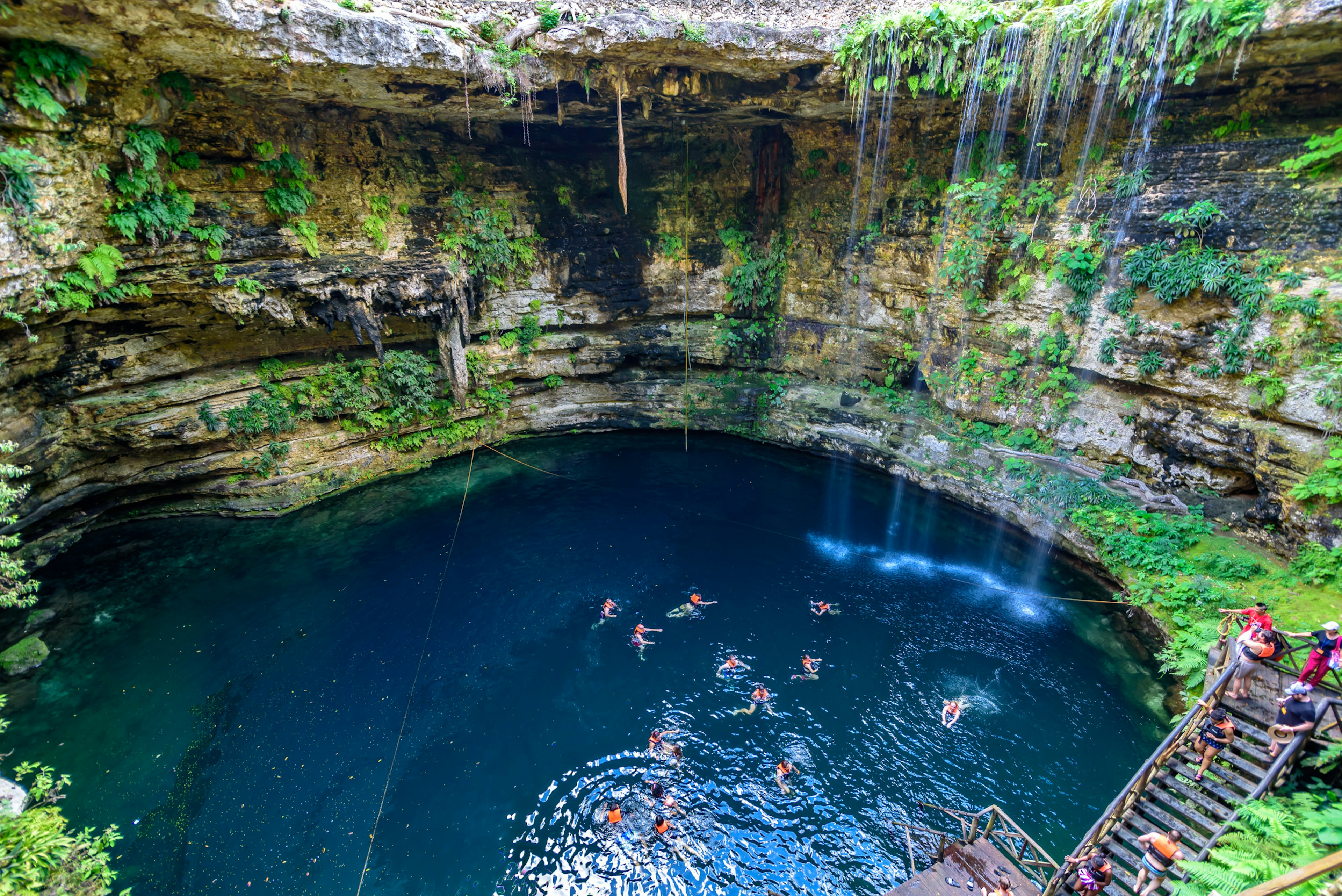 An aerial view of people swimming in a cenote swimming hole. Greenery covers the rock face, with water pouring over one side. Other swimmers descend a flight of stairs toward the water.