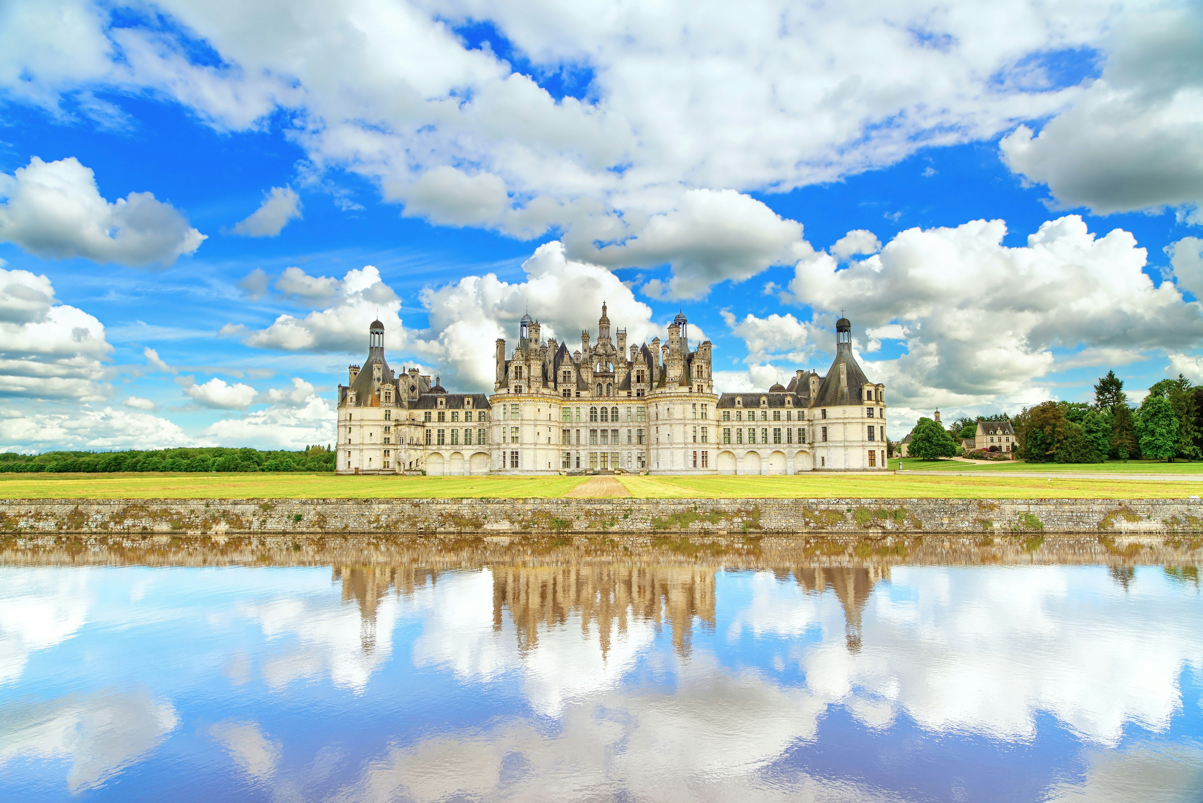 Chateau de Chambord, an impressive white-brick renaissance building with turrets and a grey slate roof. There is a large body of still water in front of the castle that perfectly mirrors the cloudy blue sky above